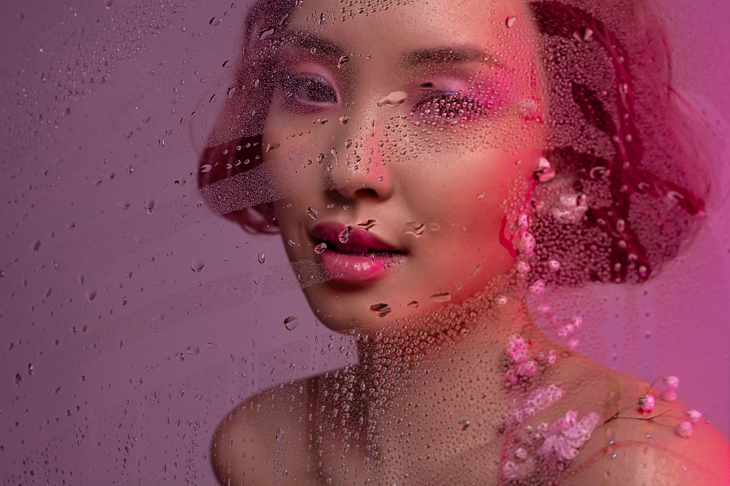 Задумчивый портрет юной азиатки с цветочными акцентами на руках и шее, ее взор направлен на капли воды, стекающие по стеклу, предвещая пробуждение природы.