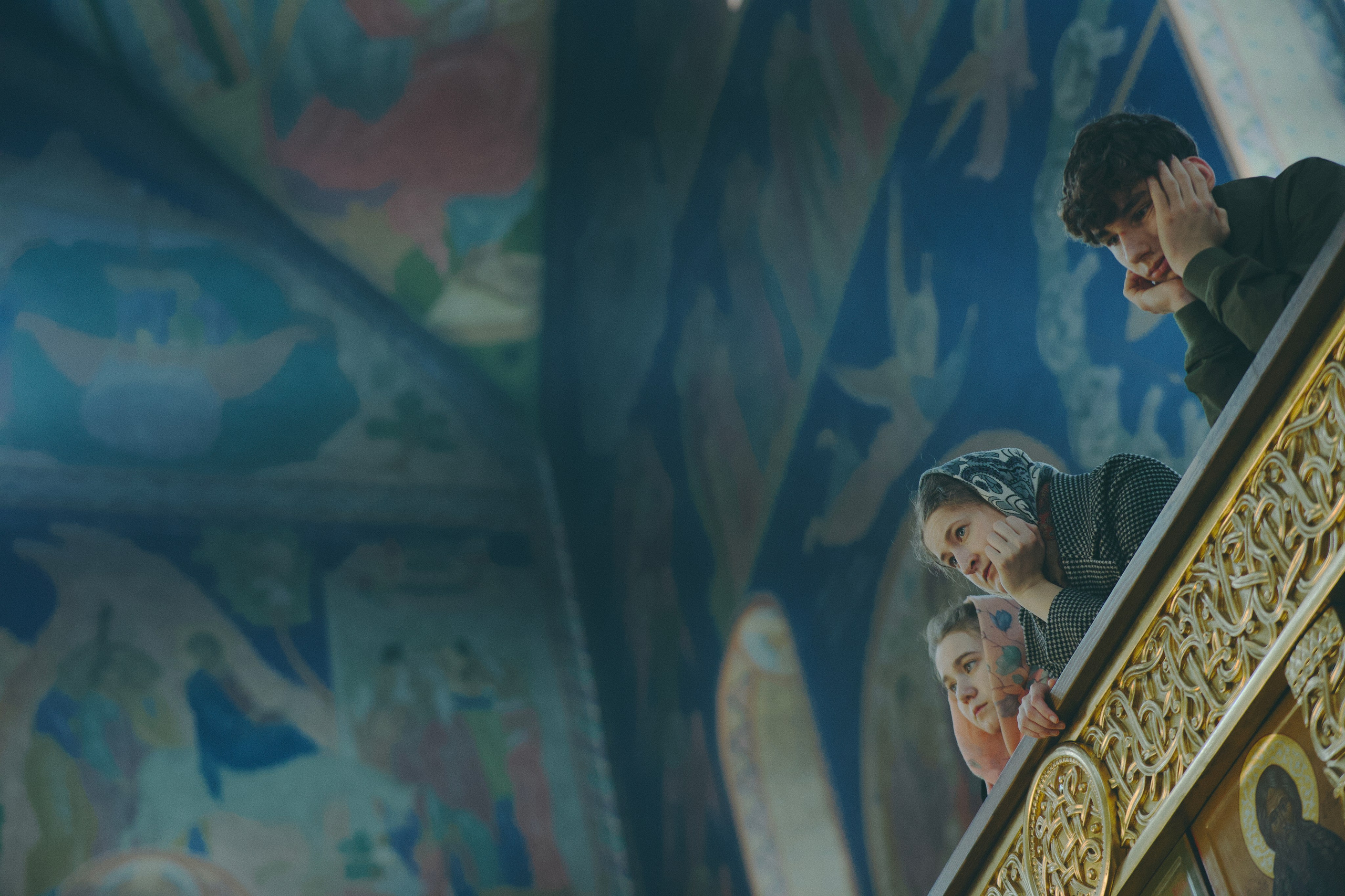 Юноша и две девушки с платками смотрят вниз с клироса/балкона на богослужение. За ними — яркие фрески с ангелами на потолке.