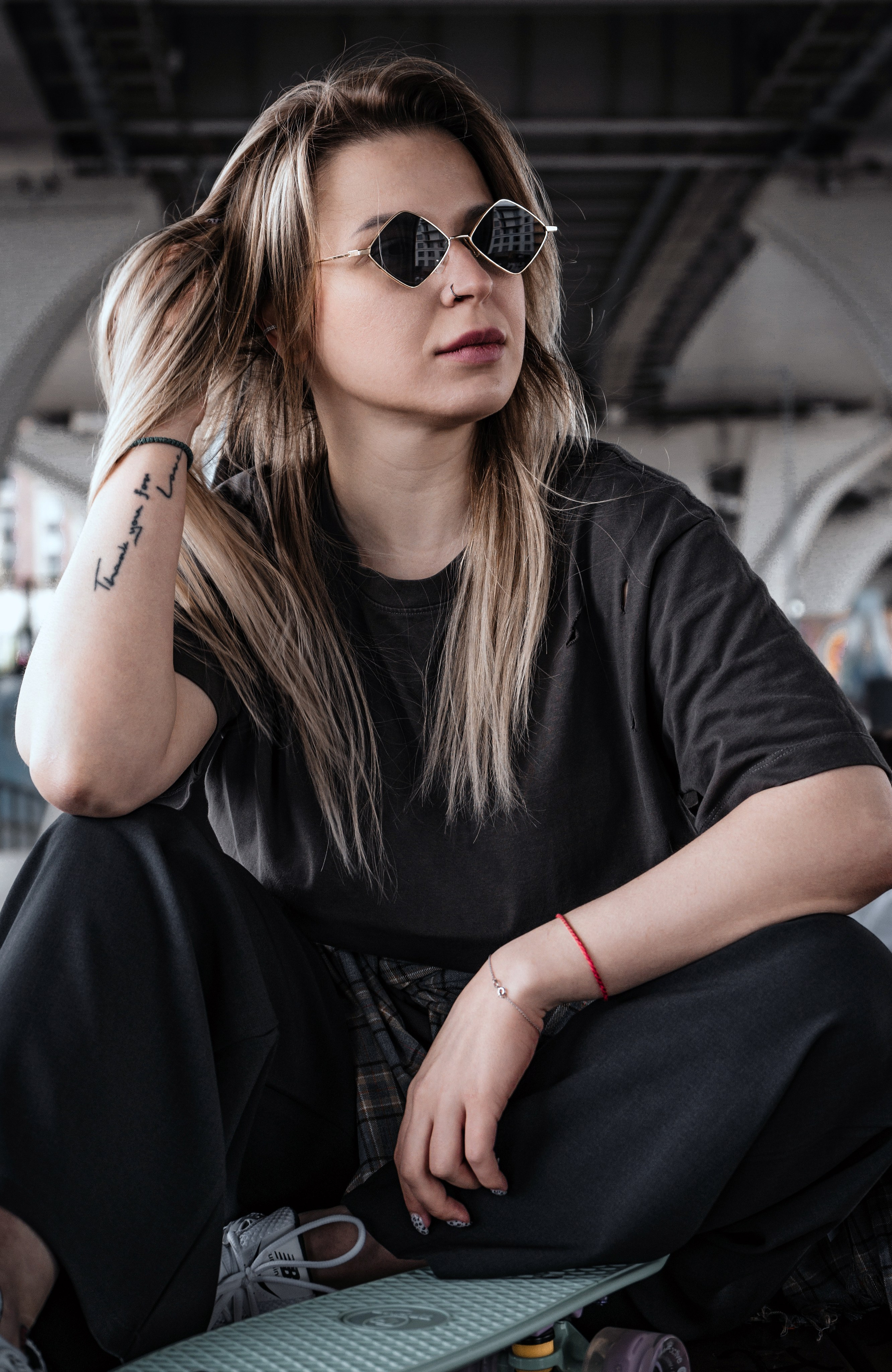 Уличная скейтер-девушка в oversize чёрной футболке и baggy брюках сидит на мятном круизёре, поправляет волосы, золотые cat-eye солнцезащитные очки, носовой пирсинг, татуировка на предплечье, городской бетонный фон.
