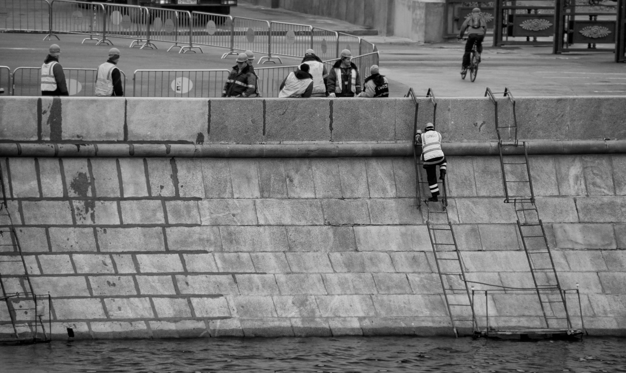 Рабочие в жилетах на каменной набережной с лестницами, велосипедист на фоне, черно-белая уличная фотография