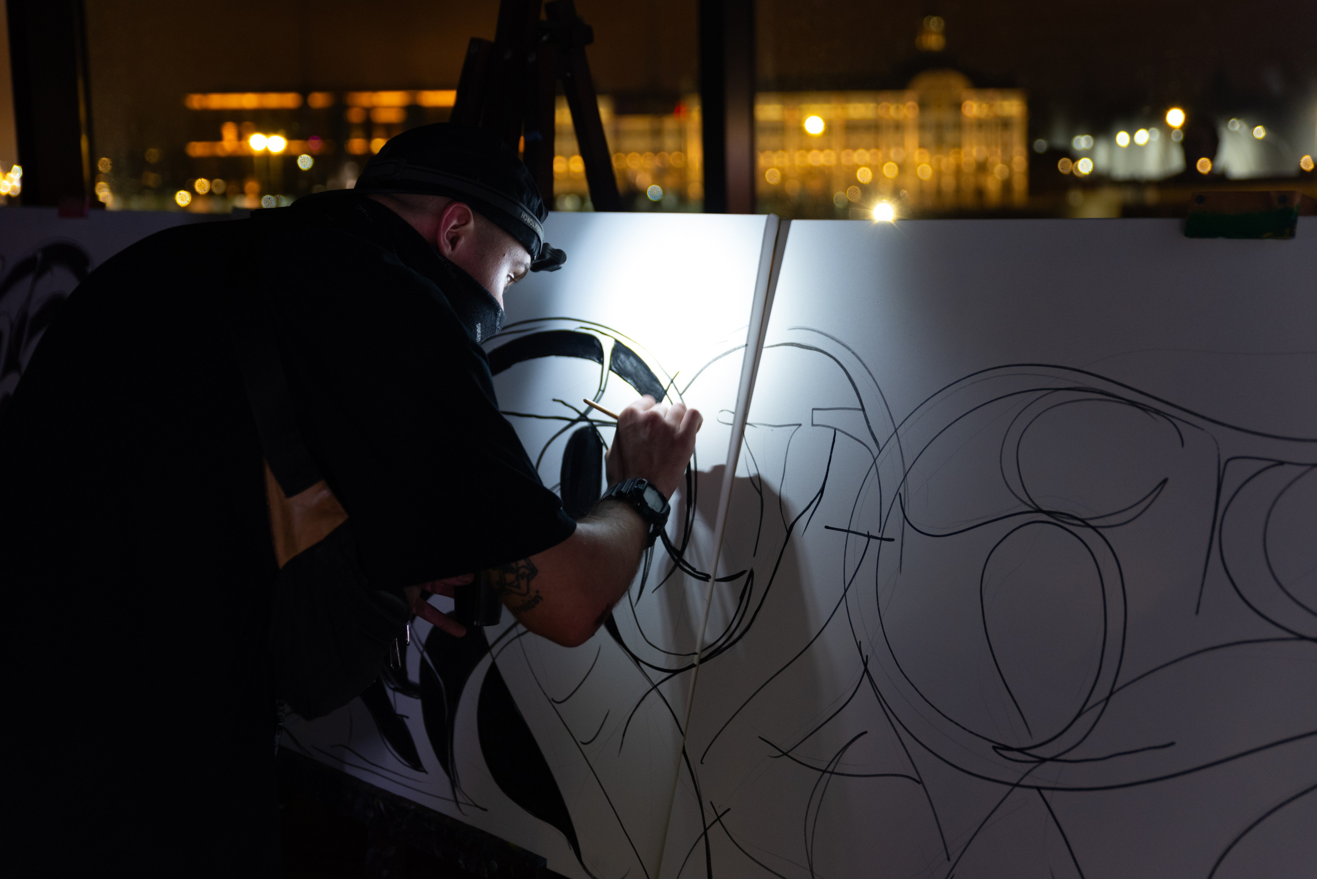 Художник рисует графический узор на белом холсте, вокруг темнота, за холстом большое окно и вид на город, у художника на голове налобный фонарь