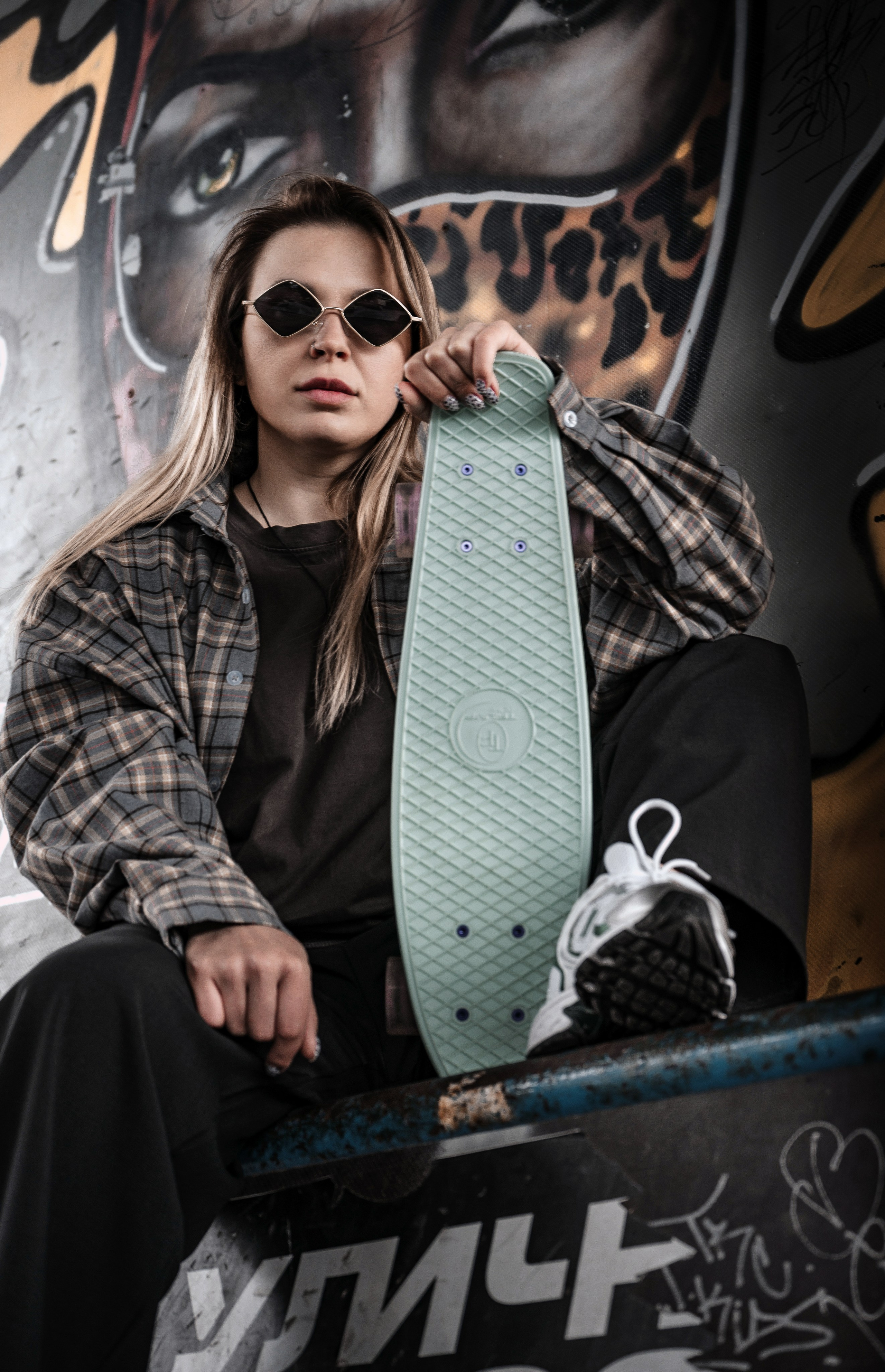 Граффити streetwear портрет — девушка с носовым пирсингом и татуировкой держит мятный круизер, oversize клетчатая рубашка, чёрная футболка, леопардовый граффити на фоне, chunky кроссовки, уличная скейт-мода.