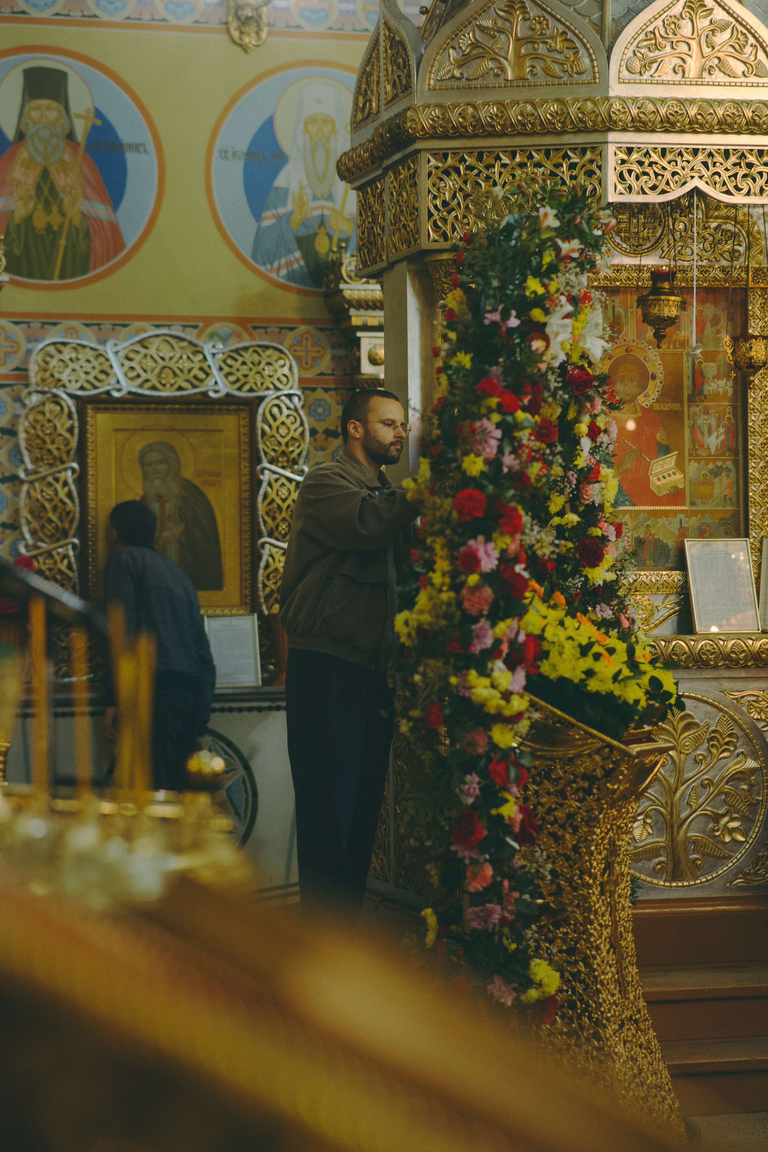 Мужчина в храме аккуратно украшает цветами высокий аналой. На заднем плане — иконостас и фрески с ликами святых.