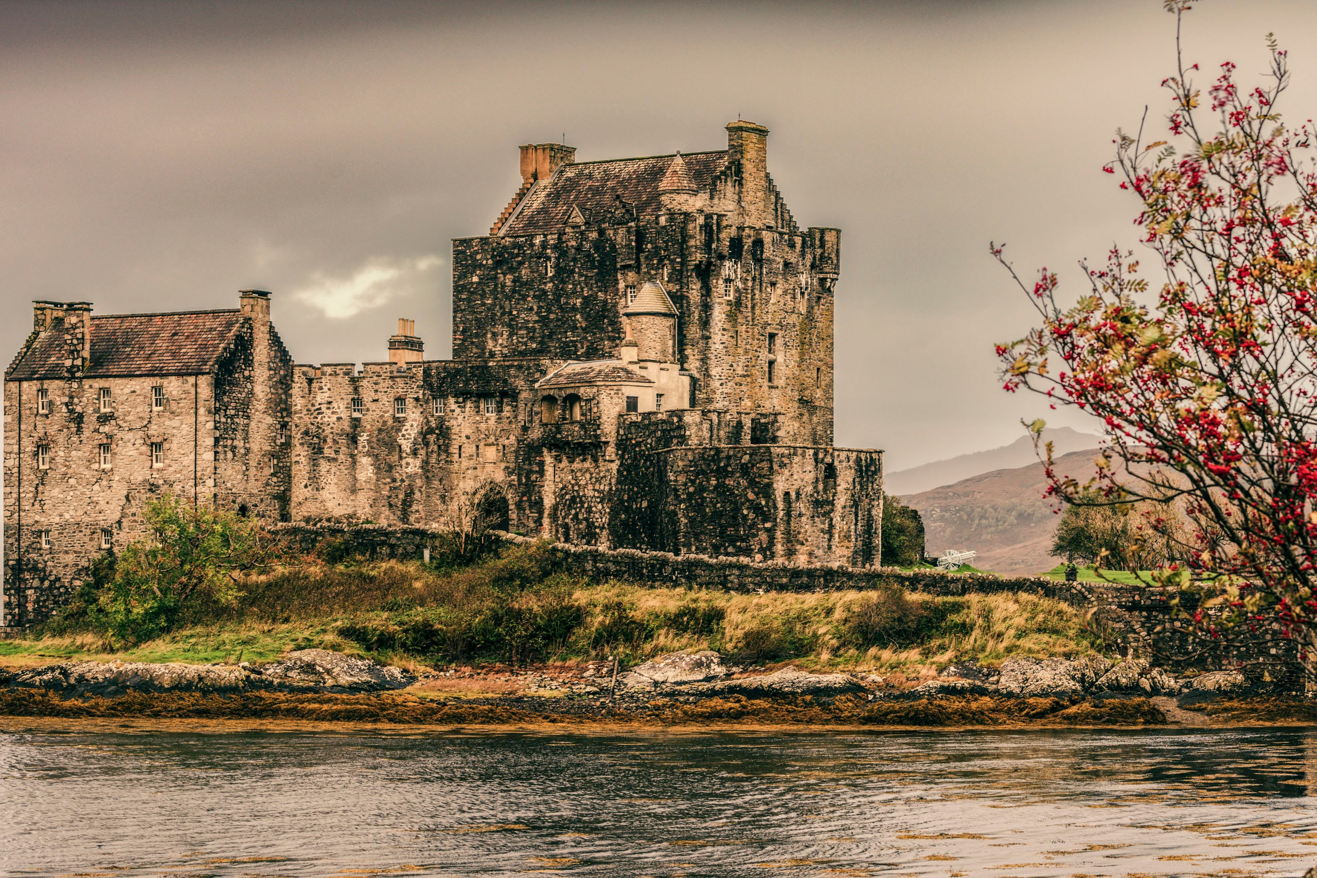 Исторический замок Эйлен Донан (Eilean Donan Castle) на небольшом острове, окруженный водой и горами Шотландского Хайленда, с осенними красными листьями дерева на переднем плане справа.