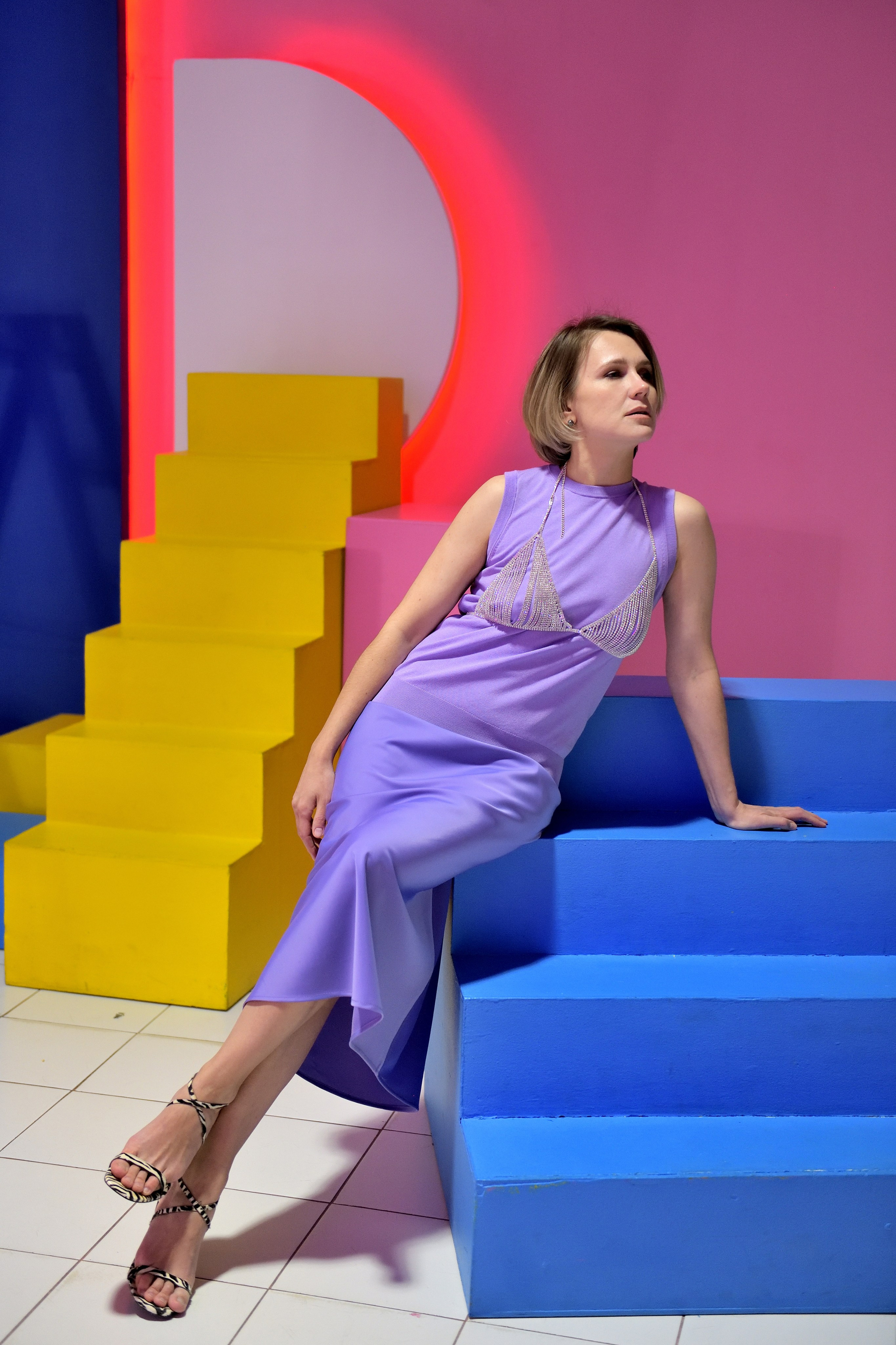 Девушка в сиреневом платье позирует на фоне цветных деталей в студии.