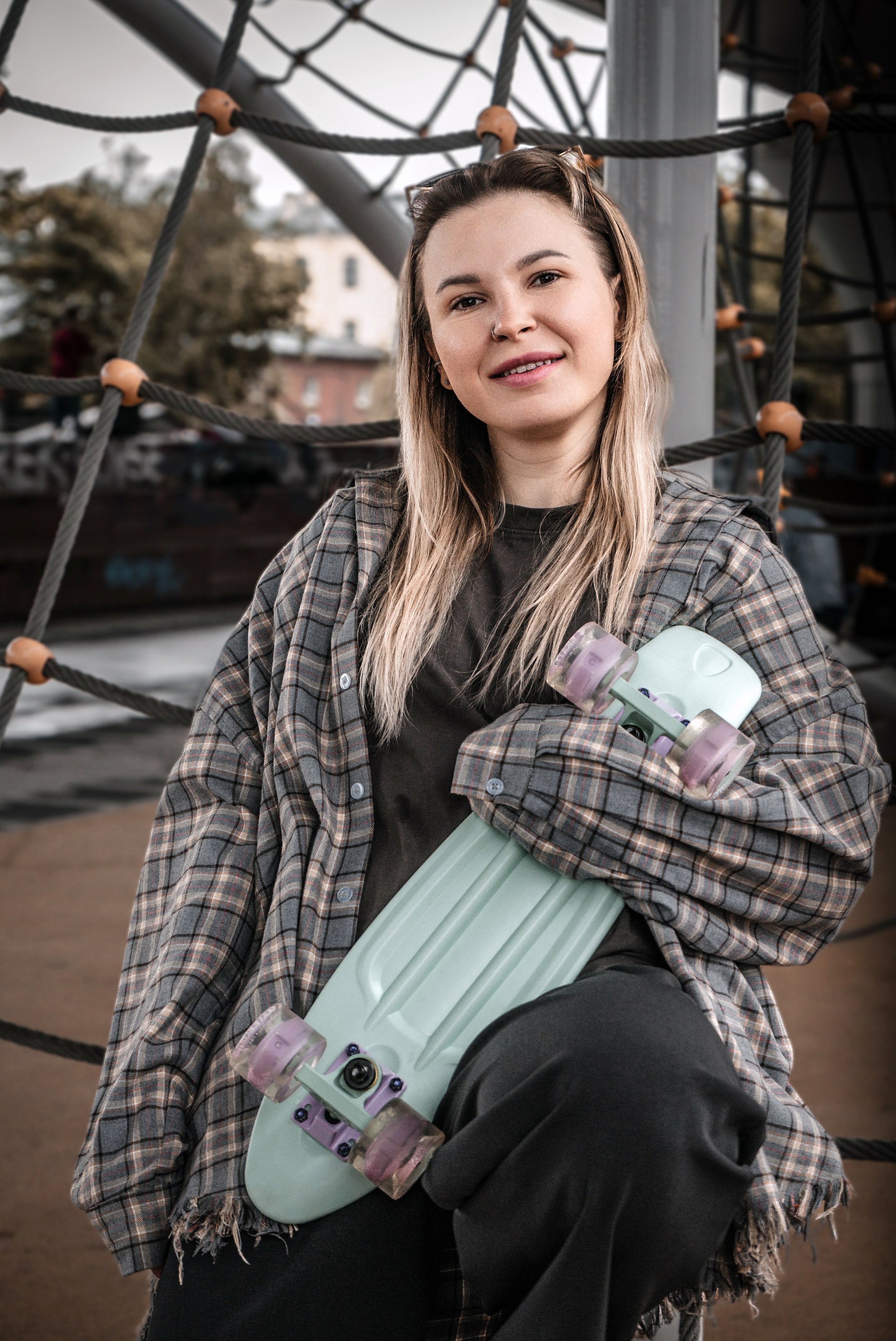 Street skater vibe: улыбающаяся девушка держит мятный penny board с фиолетовыми колёсами, oversize клетчатая рубашка в клетку поверх тёмной футболки, носовой пирсинг, детская площадка с канатами, уличная мода.