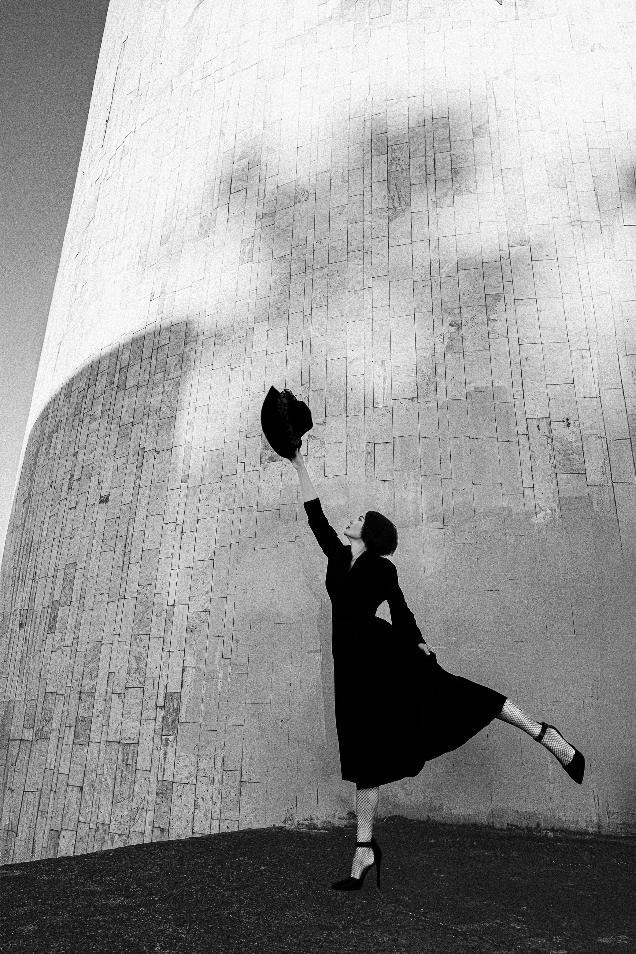 Женщина в чёрном платье и сетчатых колготках тянется вверх с шляпой на фоне высокой стены, чёрно-белая концептуальная фотосъёмка