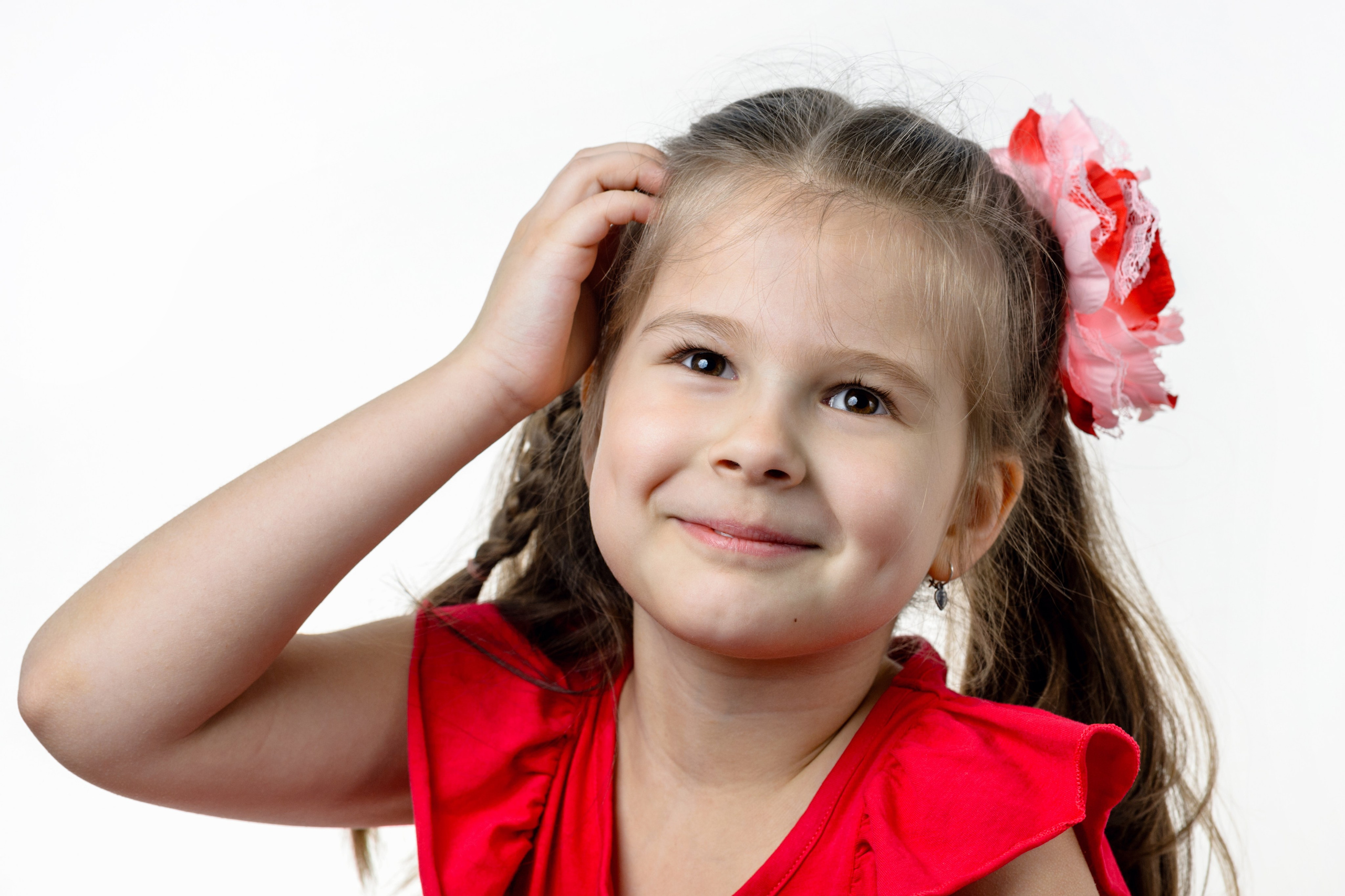 Девочка с бантиками в красном платье позирует, подняв руку к голове, на белом фоне.