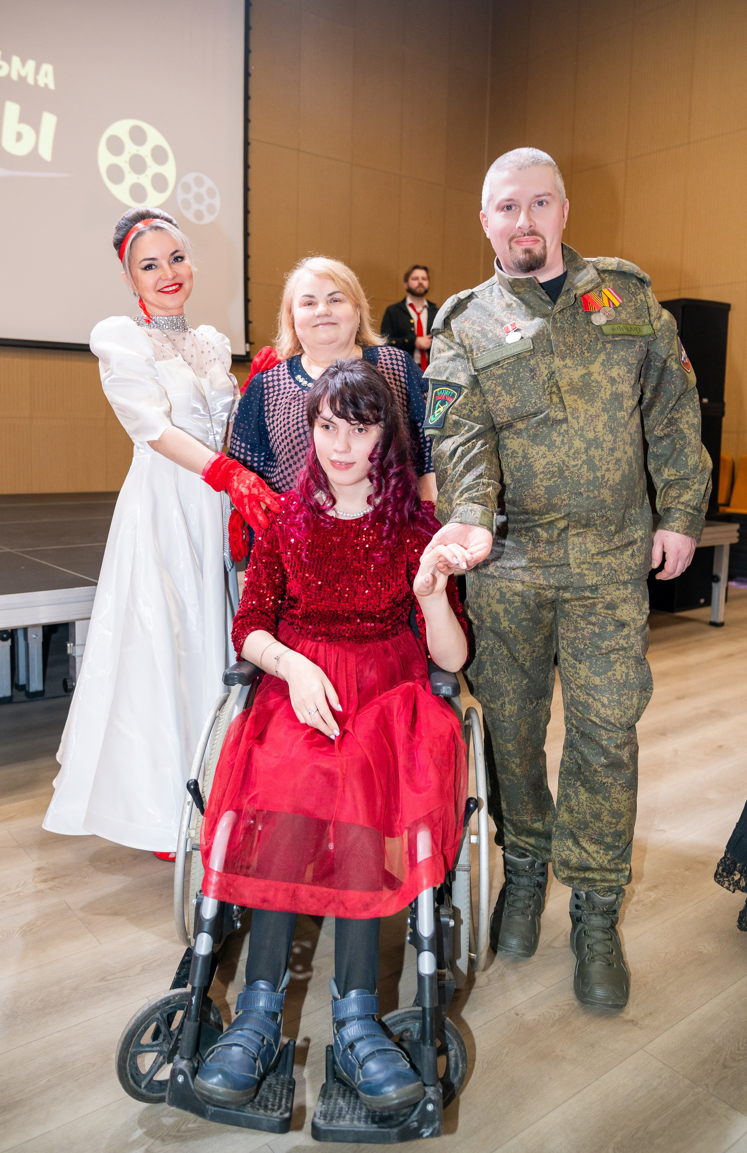 Девушка в красном блестящем платье на инвалидной коляске с мужчиной в военной форме и женщиной в белом платье на инклюзивном балу. Фотограф Мальцев Евгений.