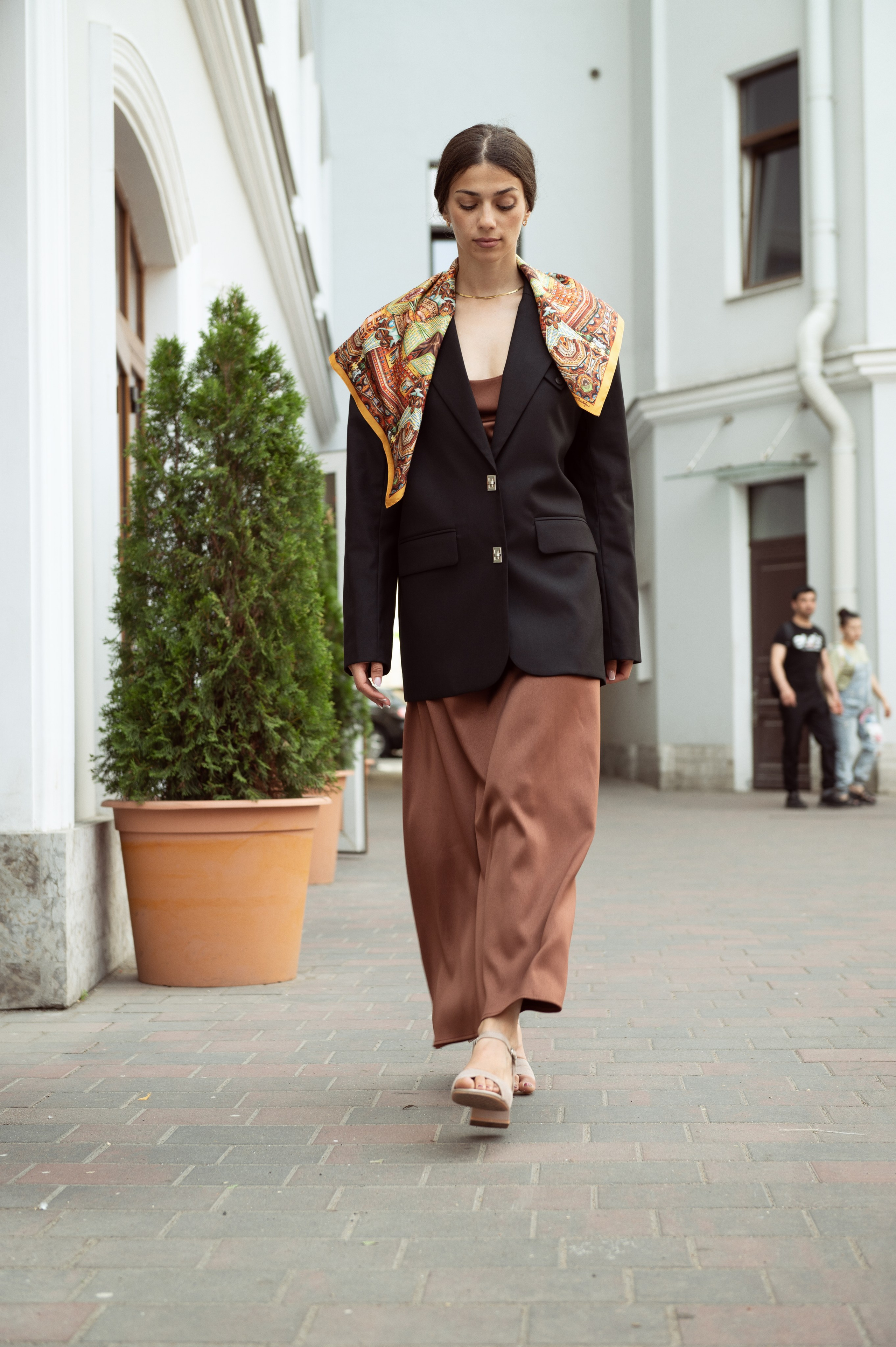 Красивая темноволосая девушка в черном пиджаке и терракотовом платье-комбинации идет по улице, у нее на плечах шелковый платок от Serafim "Cristofari Спас-на-крови"