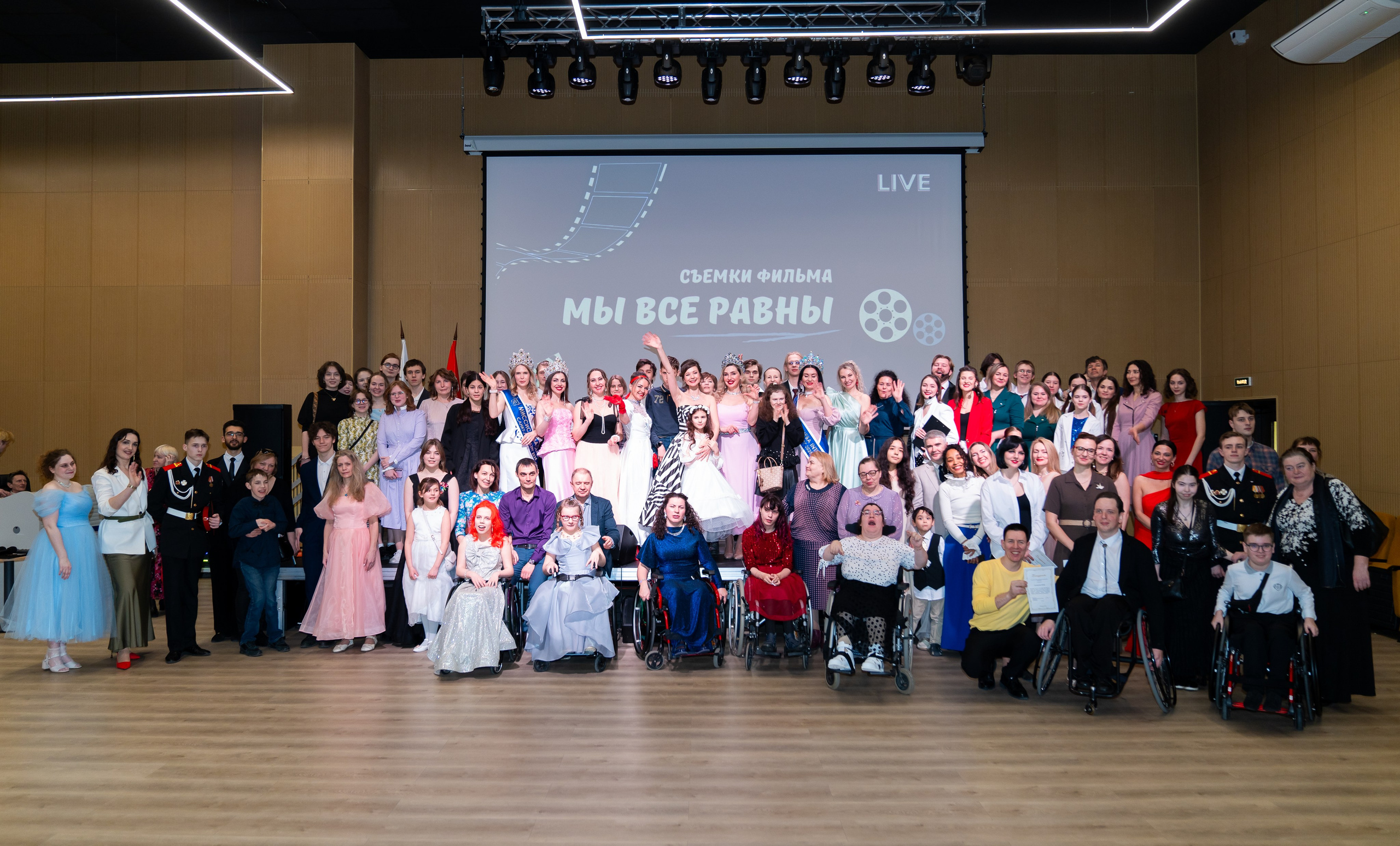 Финальное фото всех участников и гостей инклюзивного бала "Мы все равны" на сцене. Большая группа людей с коронами, в красивых платьях и костюмах. Фотография Евгения Мальцев.