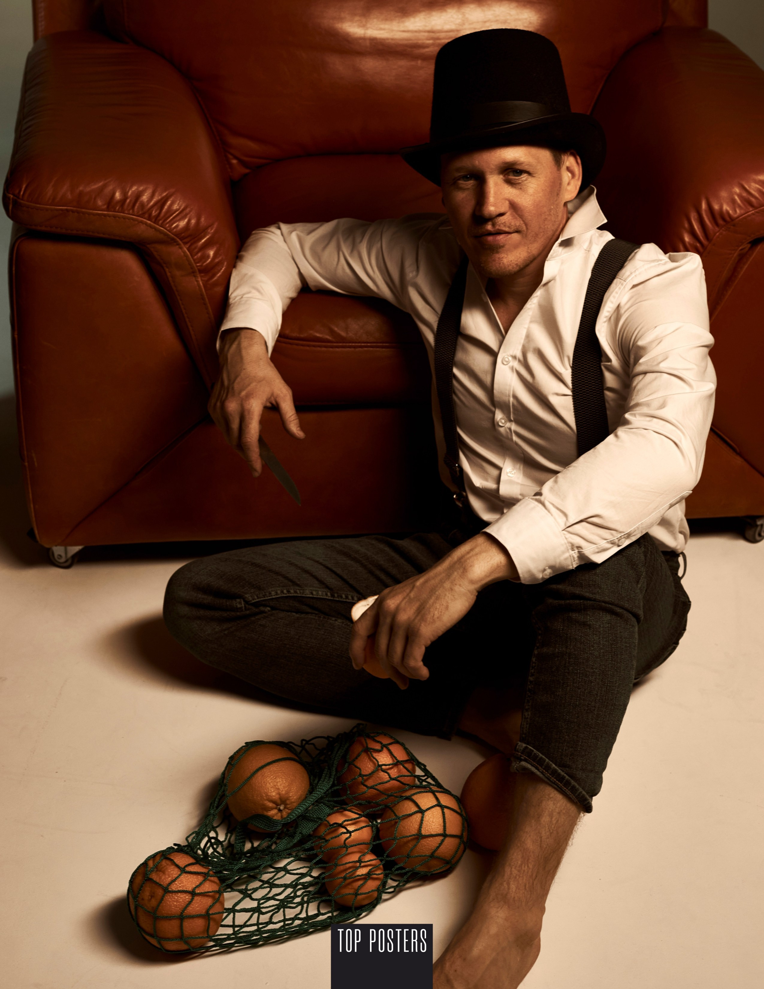 Портрет мужчины в черной шляпе-цилиндре, в белой рубашке, джинсах с подтяжками, мужчина сидит на полу и рядом авоська с апельсинами