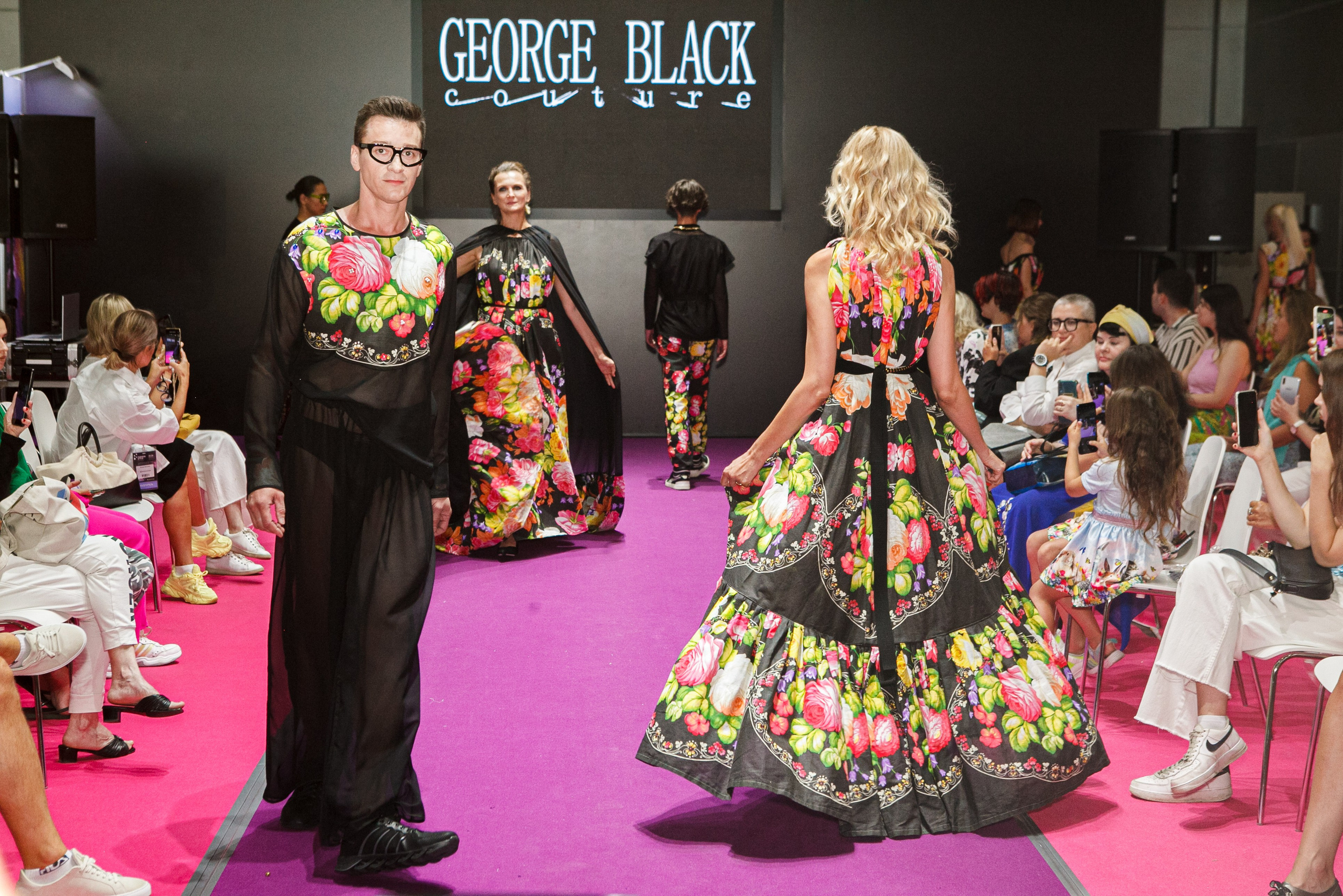  "Модели в ярких нарядах проходят по подиуму во время показа мод, освещенные яркими софитами и окруженные восторженными зрителями."