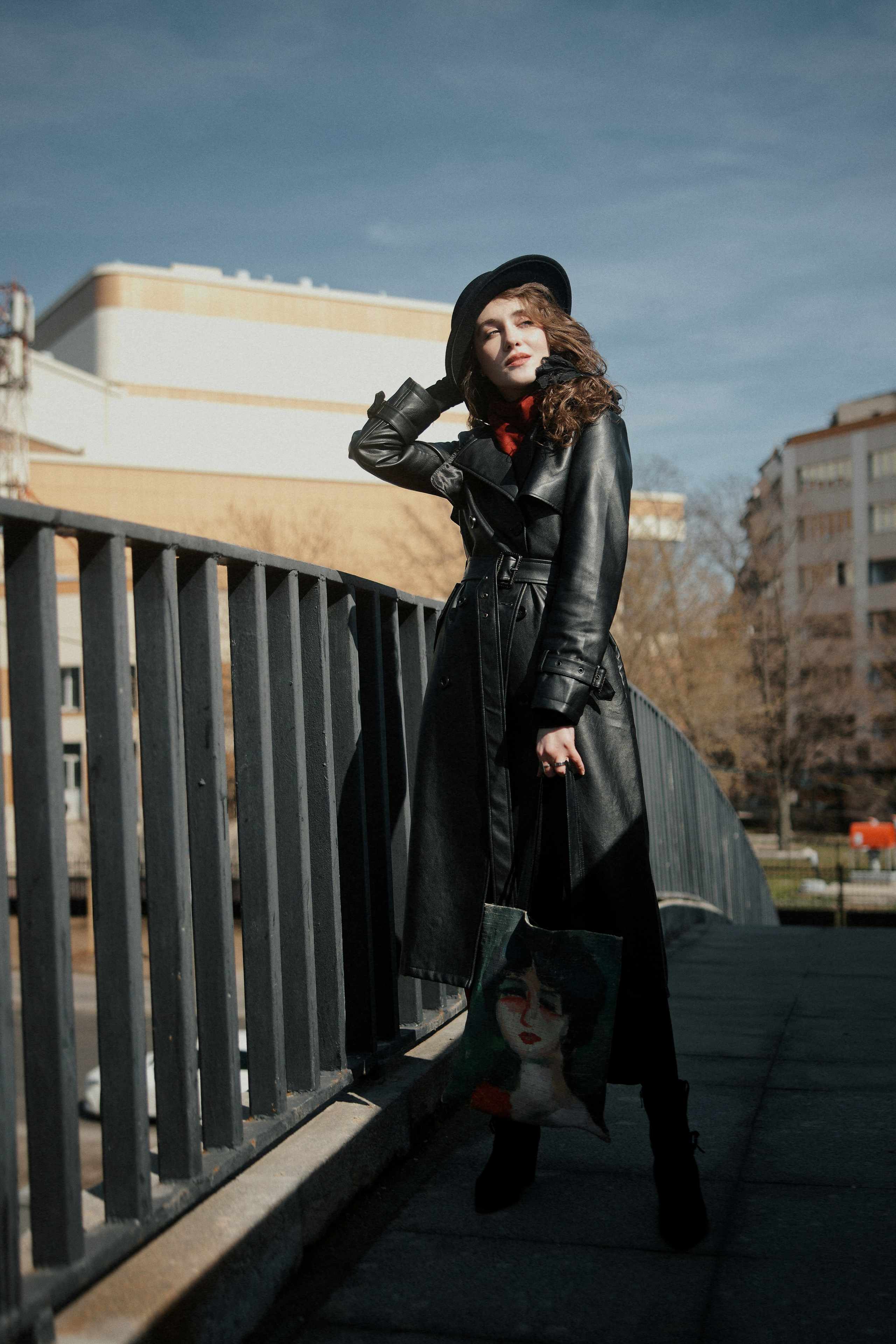 Уличный портрет девушки в длинном черном кожаном пальто и шляпе на пешеходном мосту, сумка с принтом, городской пейзаж, солнечный день