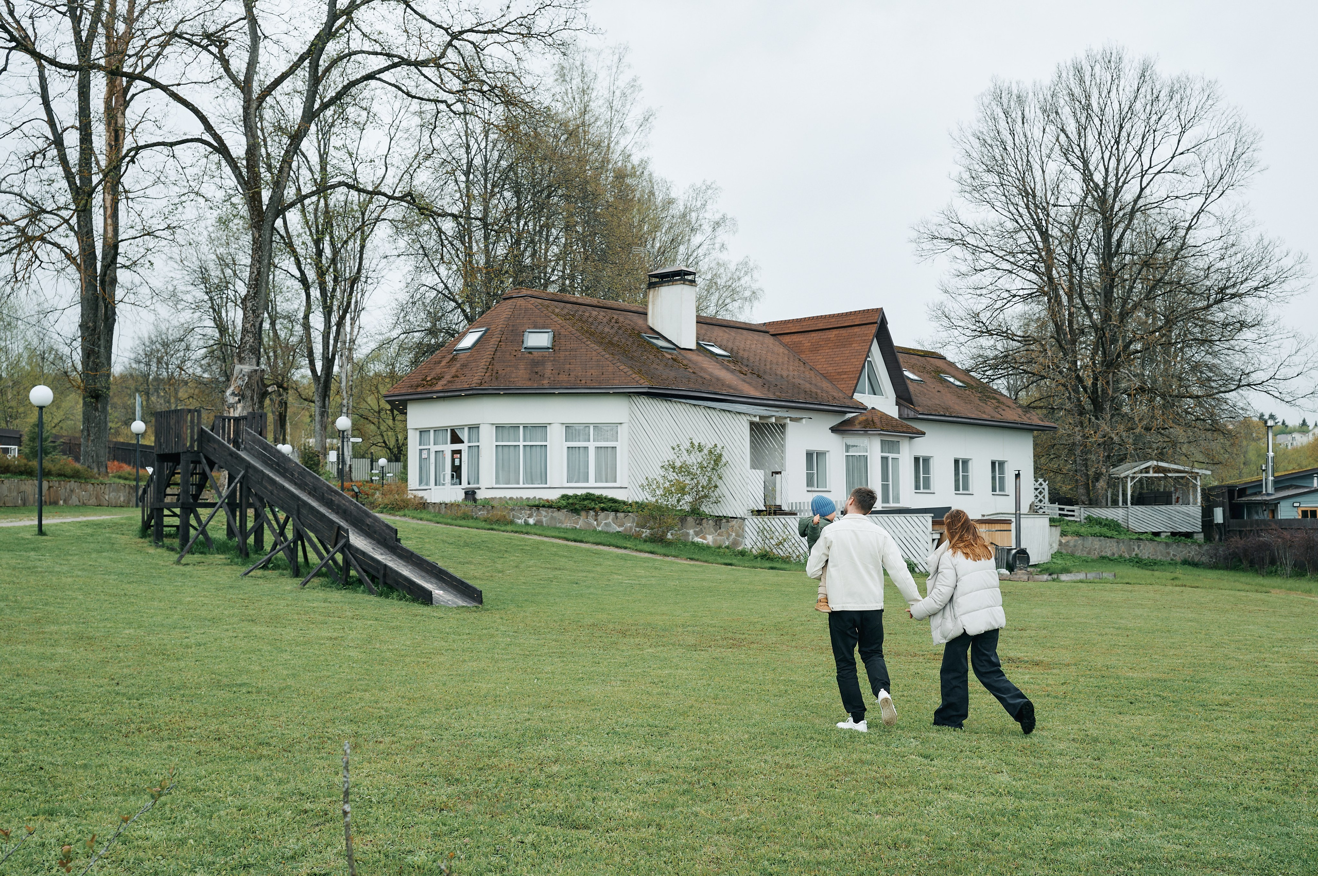 семья идёт по зелёной лужайке к белому дому с коричневой крышей