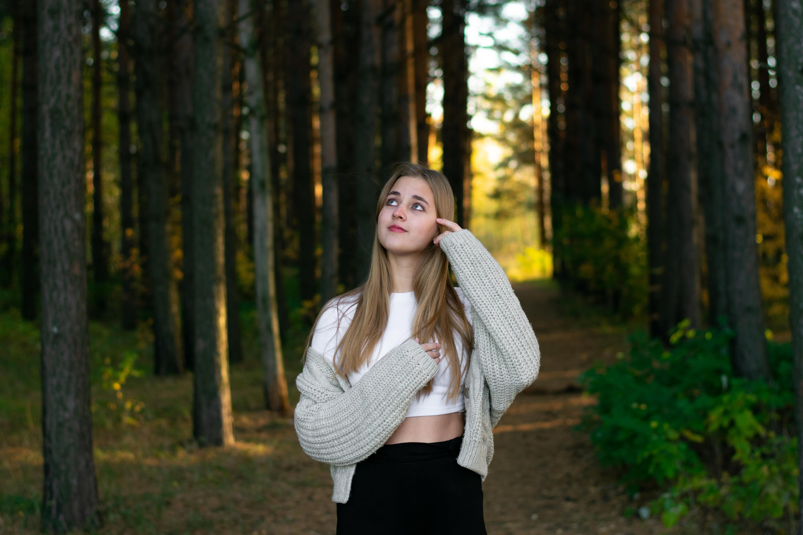 Девушка смотрит наверх в сосновом лесу в белой футболке и серой кофте