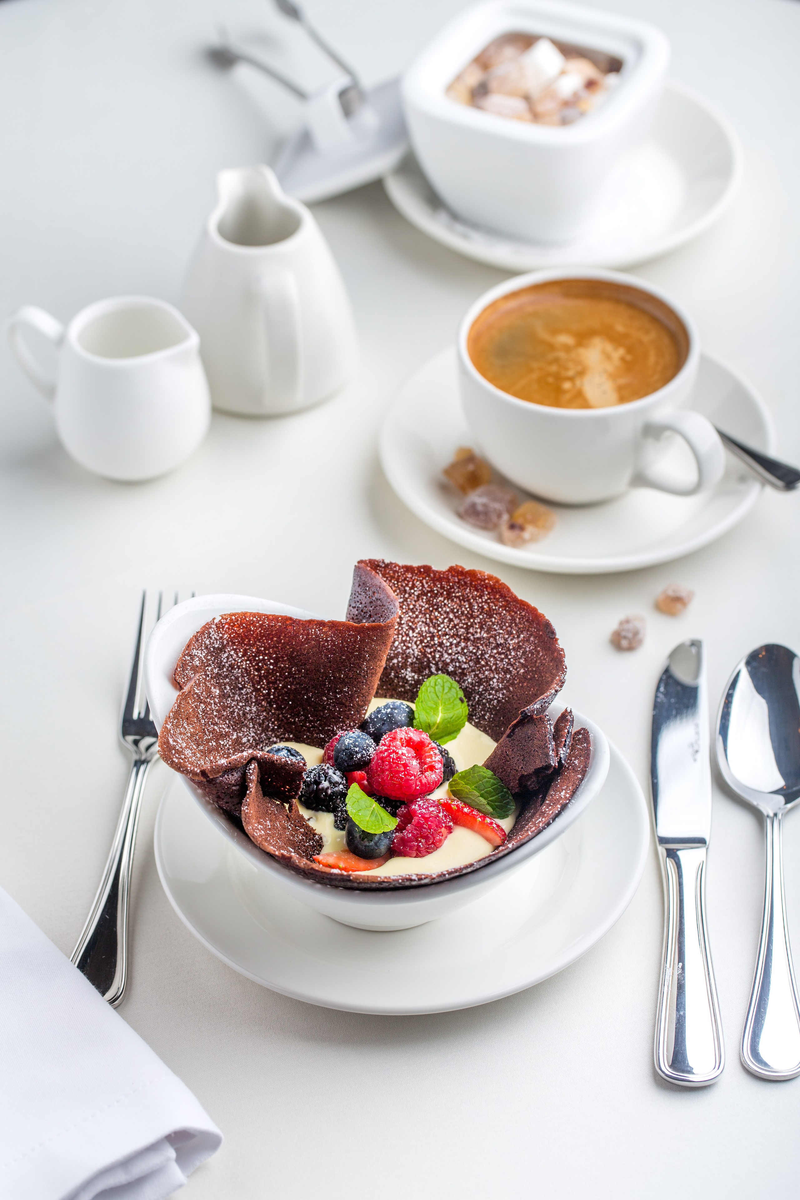 Светлая и дорогая фотокарточка десерта в белой посуде, в композиции с кофе, и приборами для создания атмосферы.