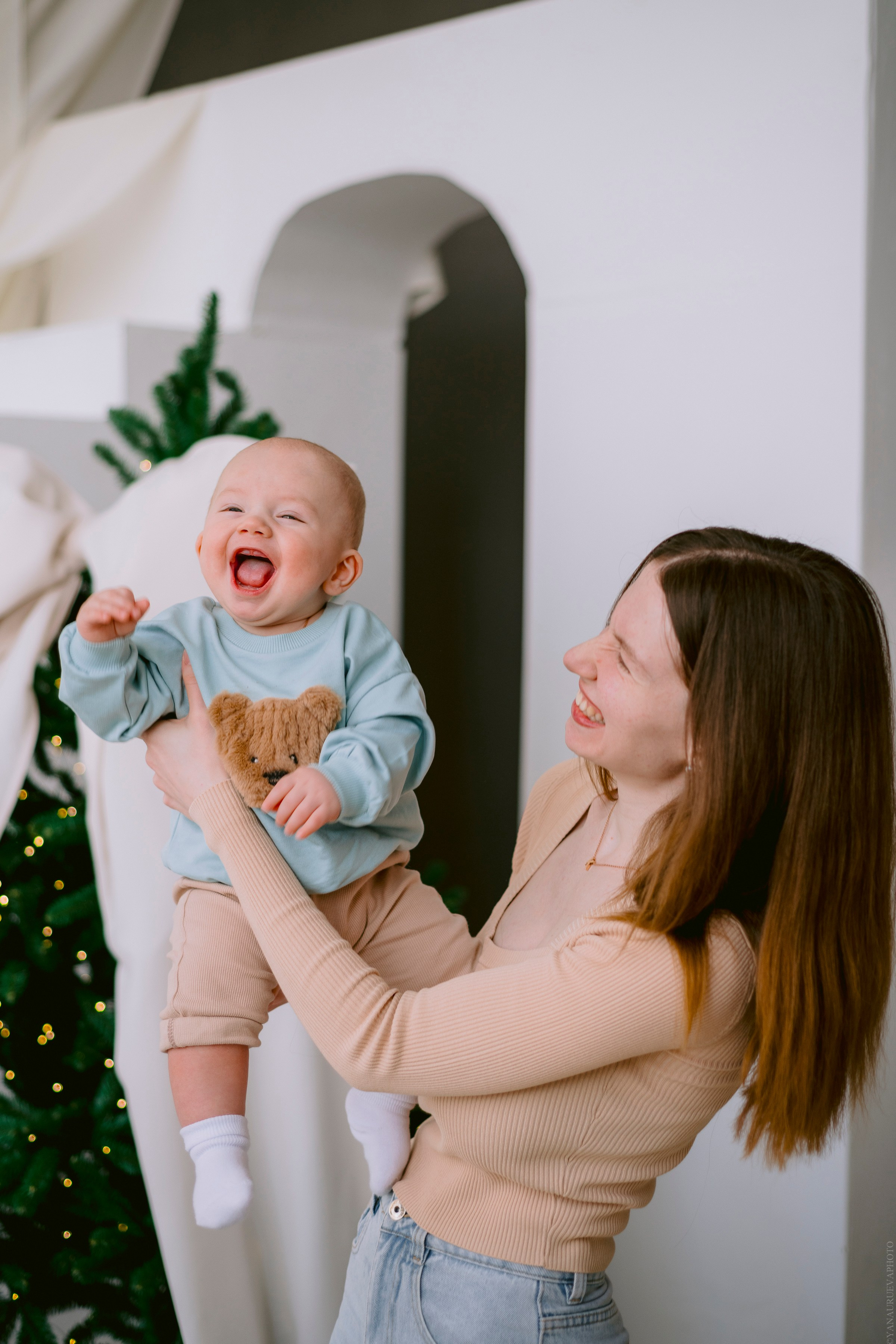 ребенок смеется на руках у мамы на фоне елки с большим бантом