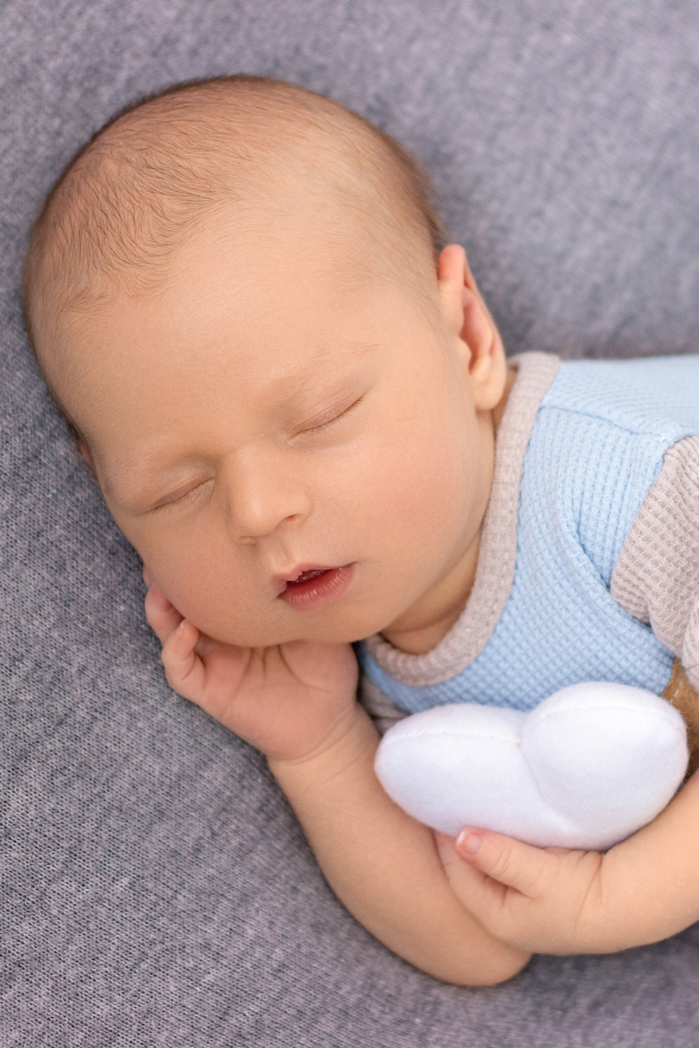 портрет новорожденного в голубо-сером костюмчике с белым сердечком в ручке на сером фоне