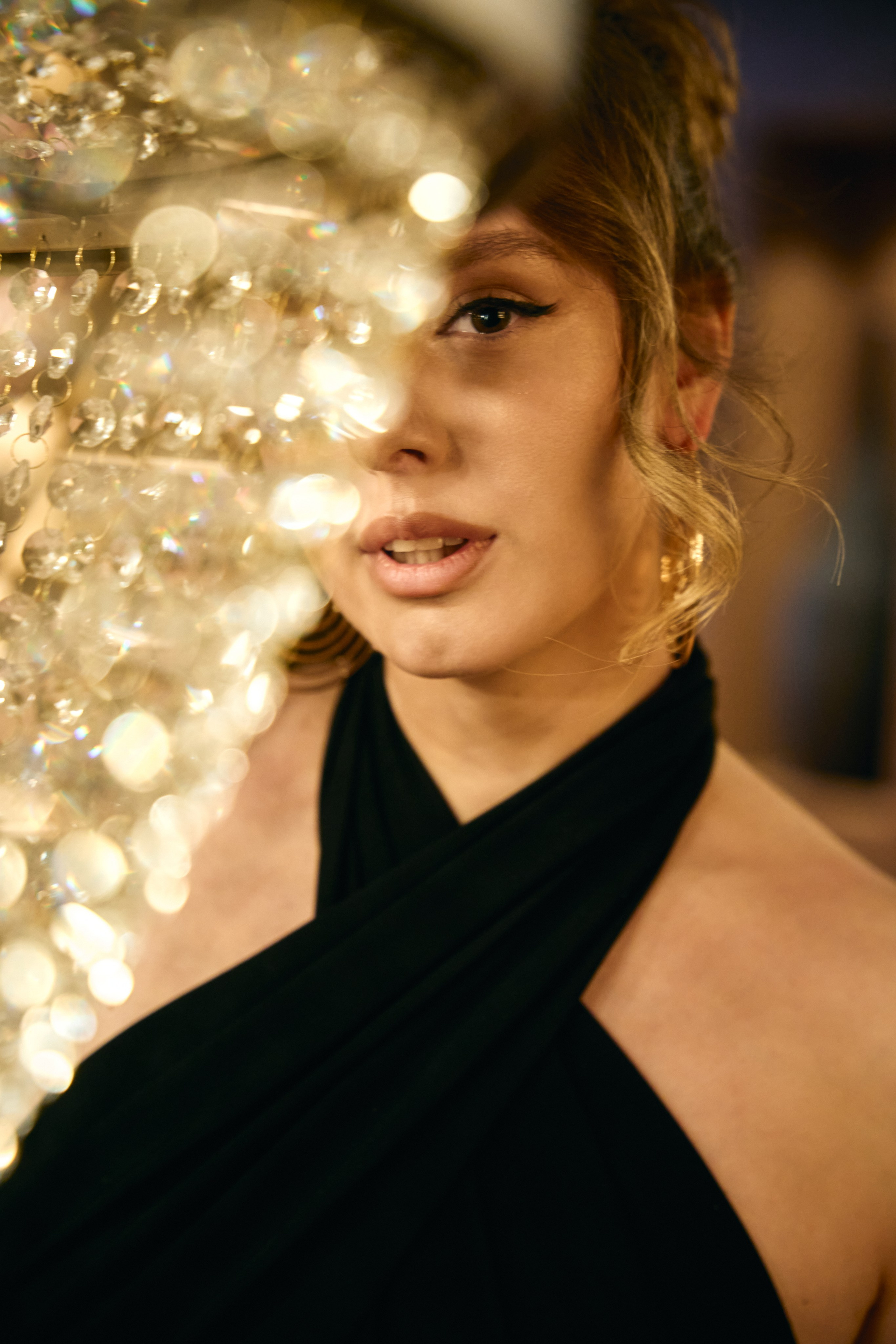 Девушка в чёрном вечернем платье стоит у стеклянной люстры в тёплом интерьере ресторана