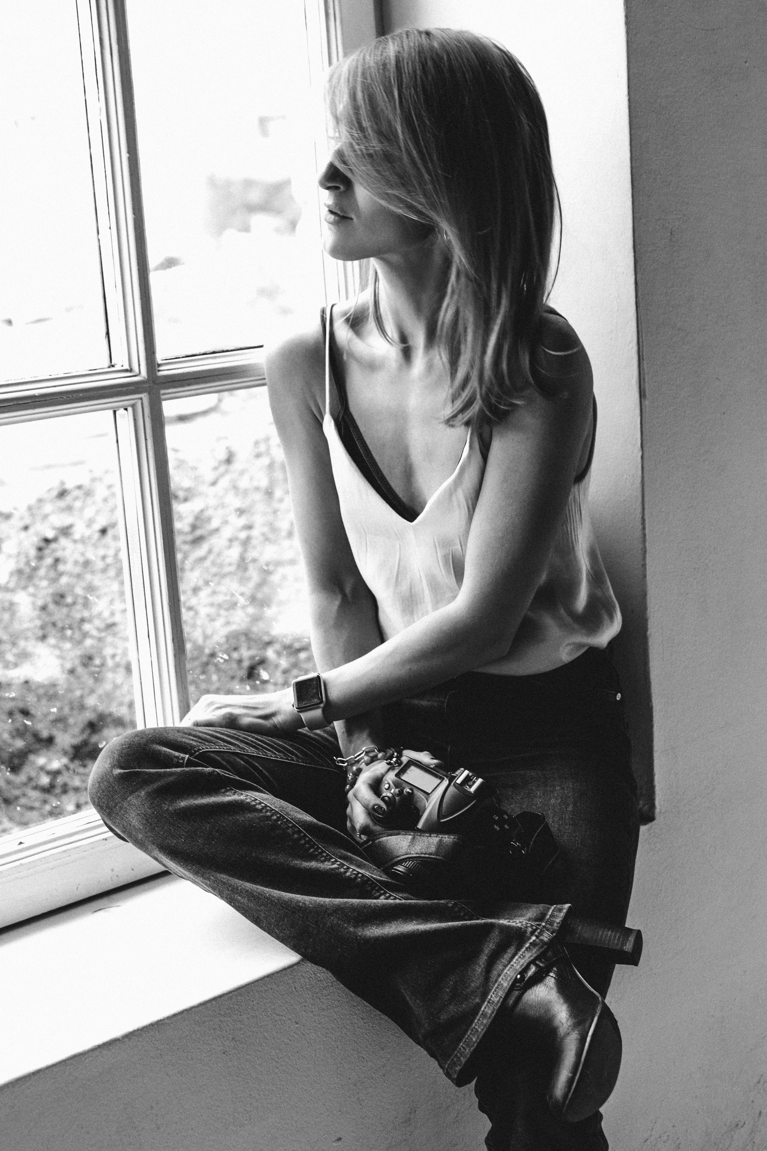 задумчивая девушка в джинсах и белой майке сидящая на подоконнике большого светлого окна