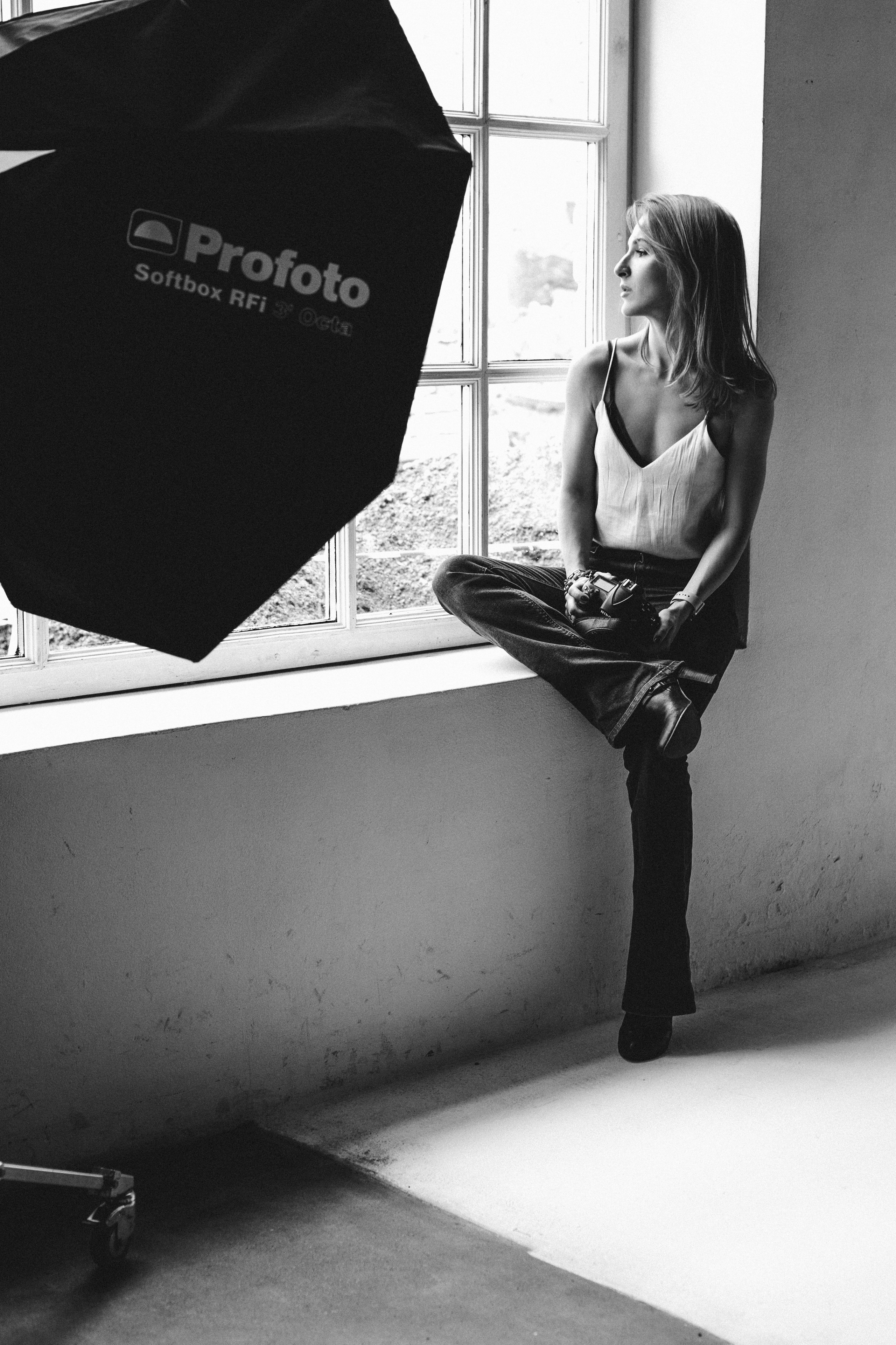 задумчивая девушка в джинсах и белой майке сидящая на подоконнике большого светлого окна фотостудии с оборудованием