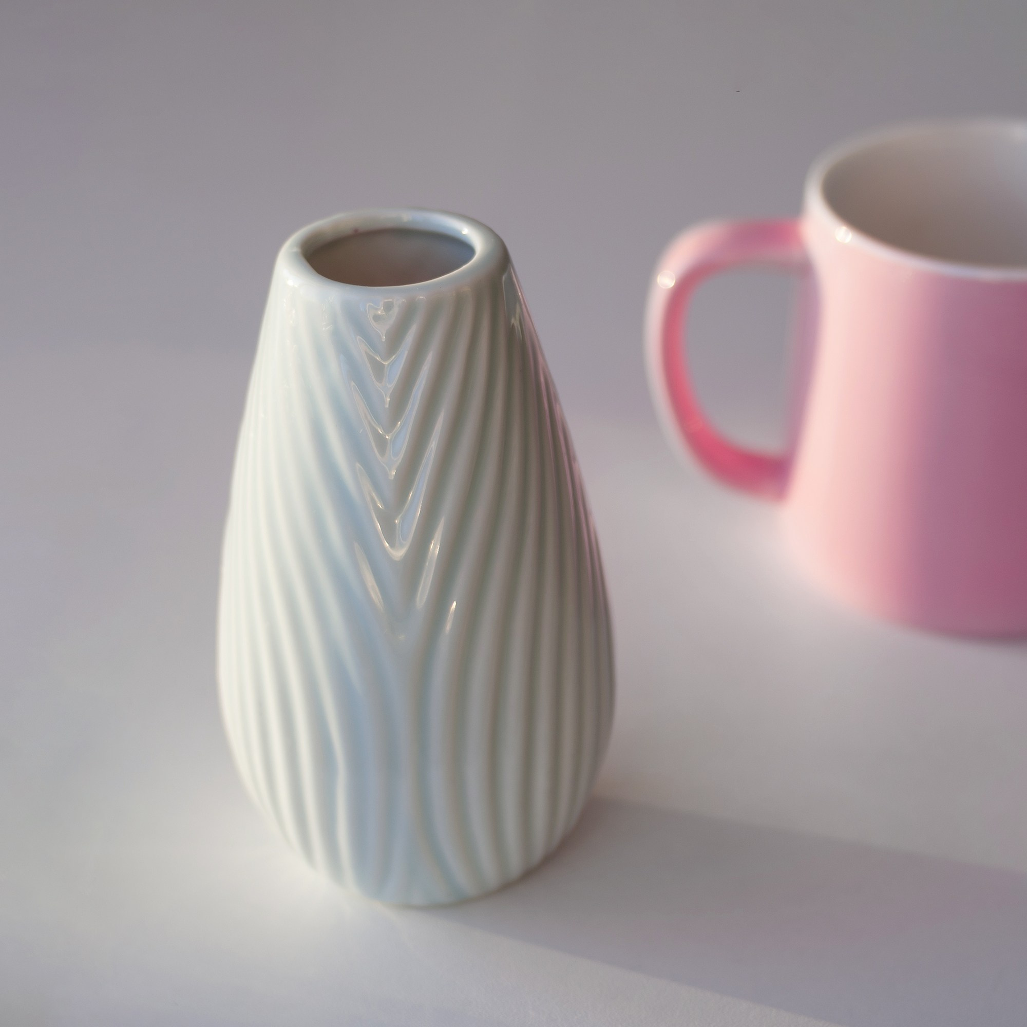 Ваза для цветов и розовая кружка на белом столе. Боковое освещение.