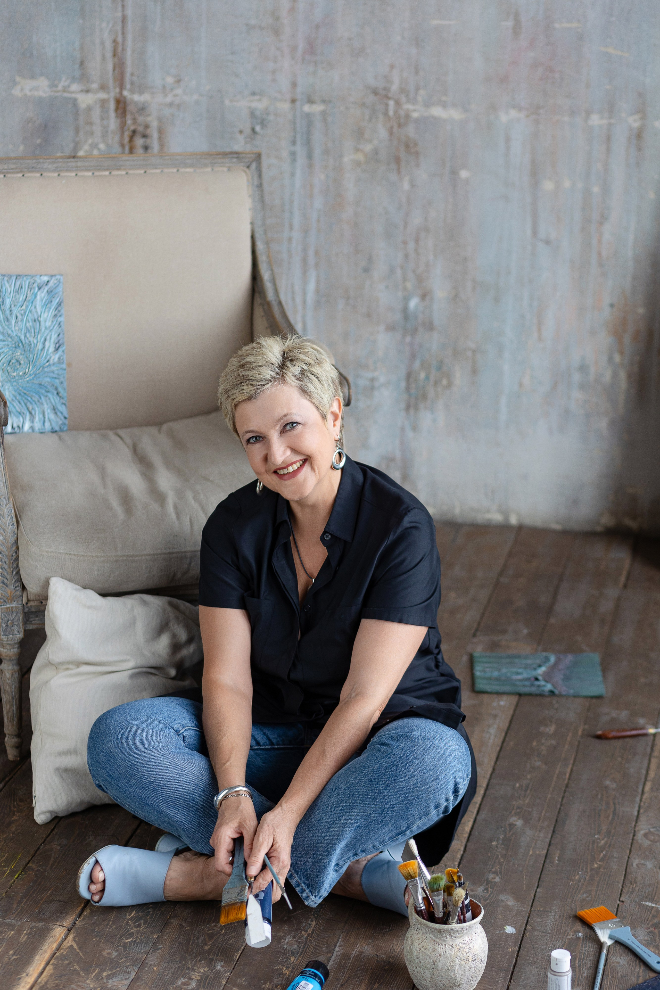 Художница сидит на полу скрестив ноги, перед ней ваза с кистями и тюбики с красками