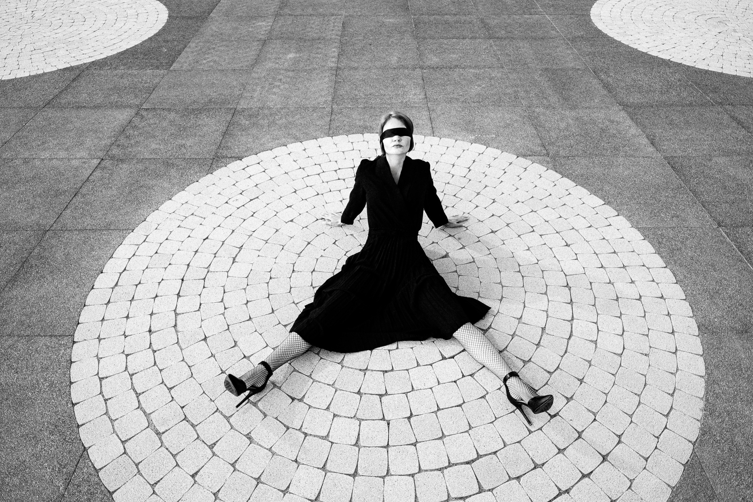 Женщина в чёрном платье и маске сидит на круглом узоре мостовой. Чёрно-белая арт-фотография с геометрическим акцентом.