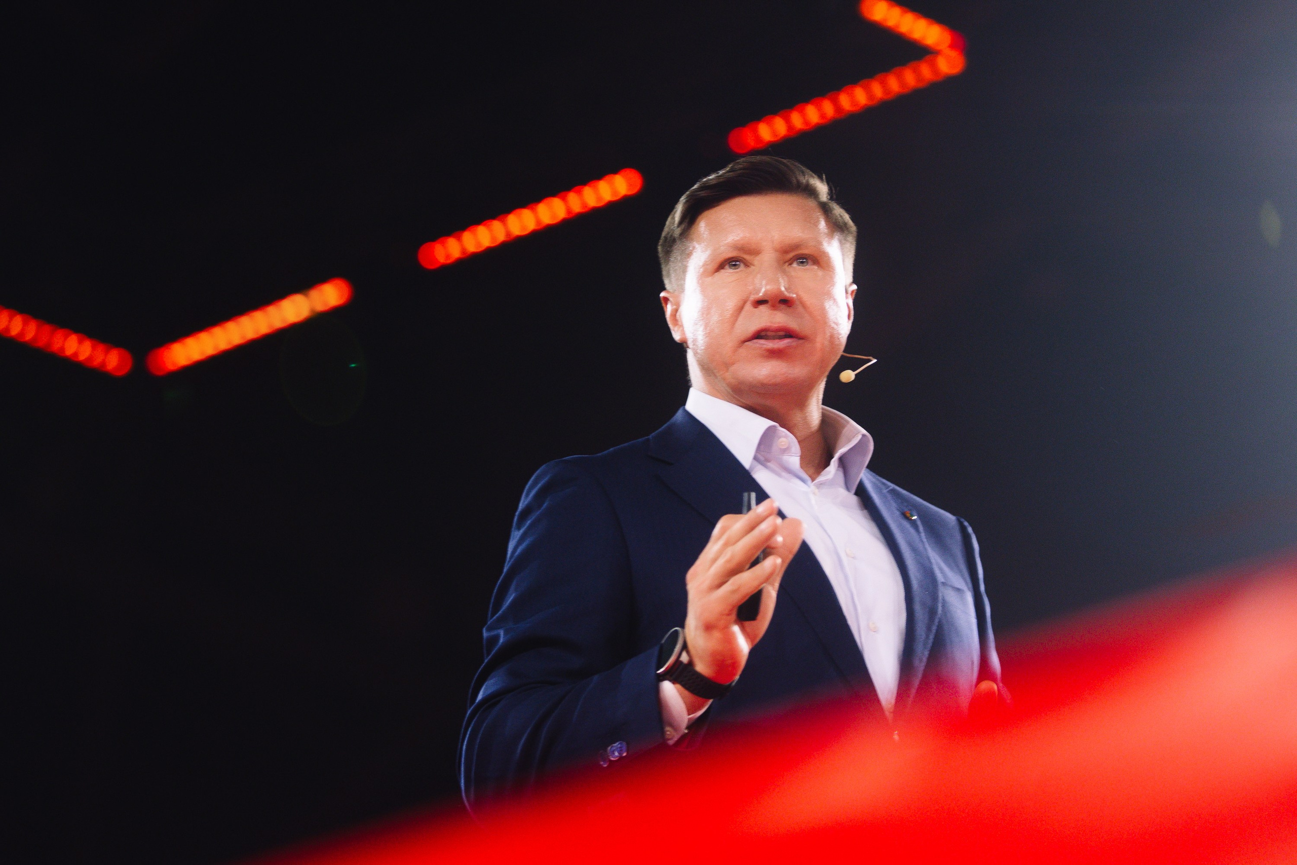 Выступление (ракурс снизу): Спикер на сцене в свете красных прожекторов, динамичный портрет лидера мнений. Владимир Волошин.