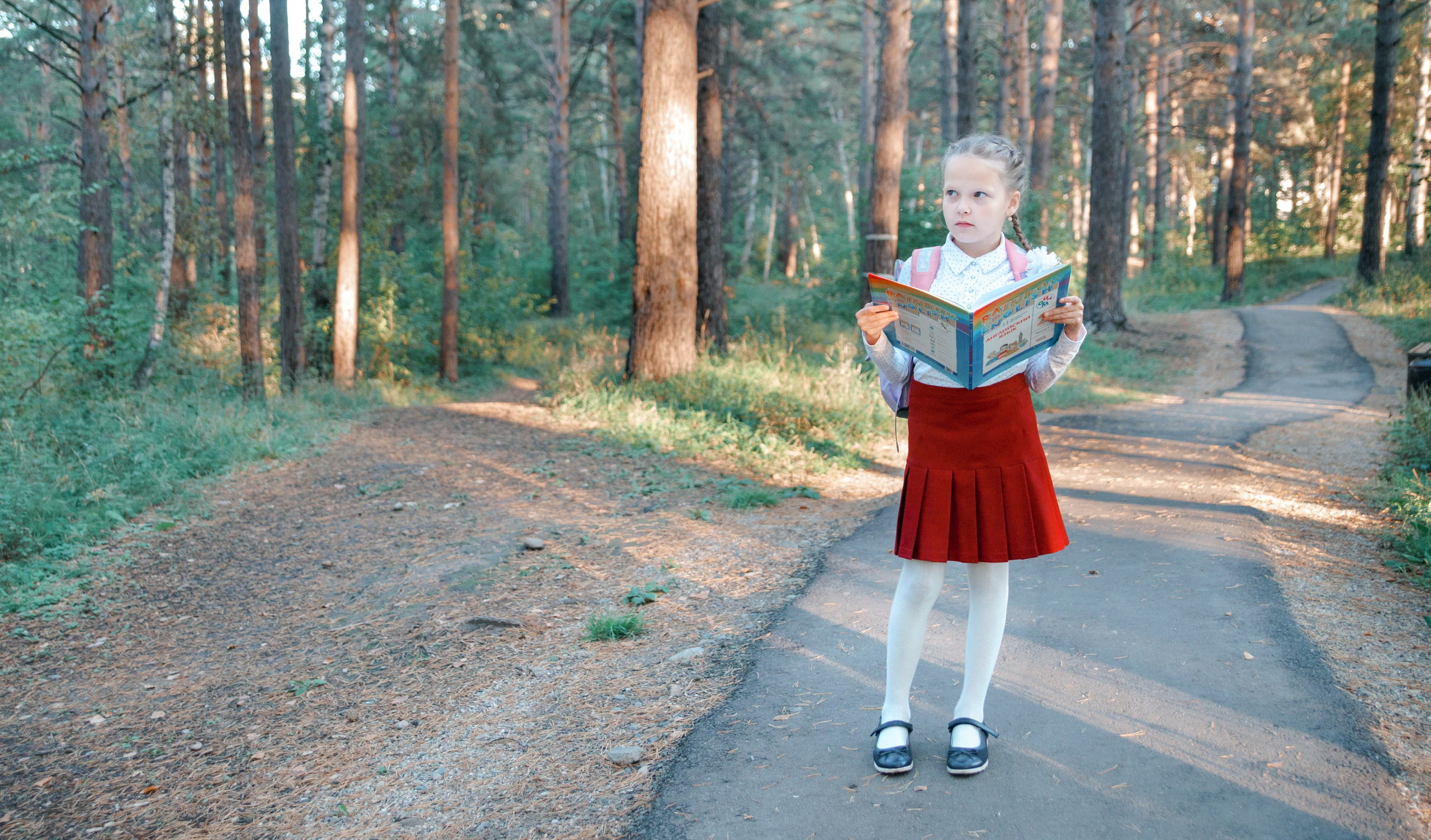 Детская фотосессия осенью - школьница стоя с учебником в лесу