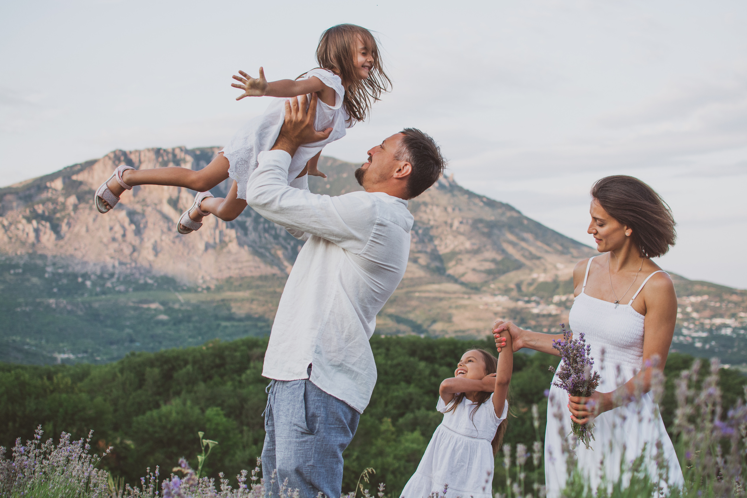 На изображении изображена семья из четырех человек, гуляющая по лавандовому полю. Родители улыбаются и держатся за руки со своими двумя маленькими детьми. На заднем плане садится солнце, а небо теплого золотистого цвета. Семья смеется и выглядит счастливой и расслабленной. Маленькую девочку в белом поднимает в воздух ее отец. Ее мать идет рядом со своим мужем, держа за руки их другую дочь. Они окружены растениями лаванды, а на заднем плане видны горы.