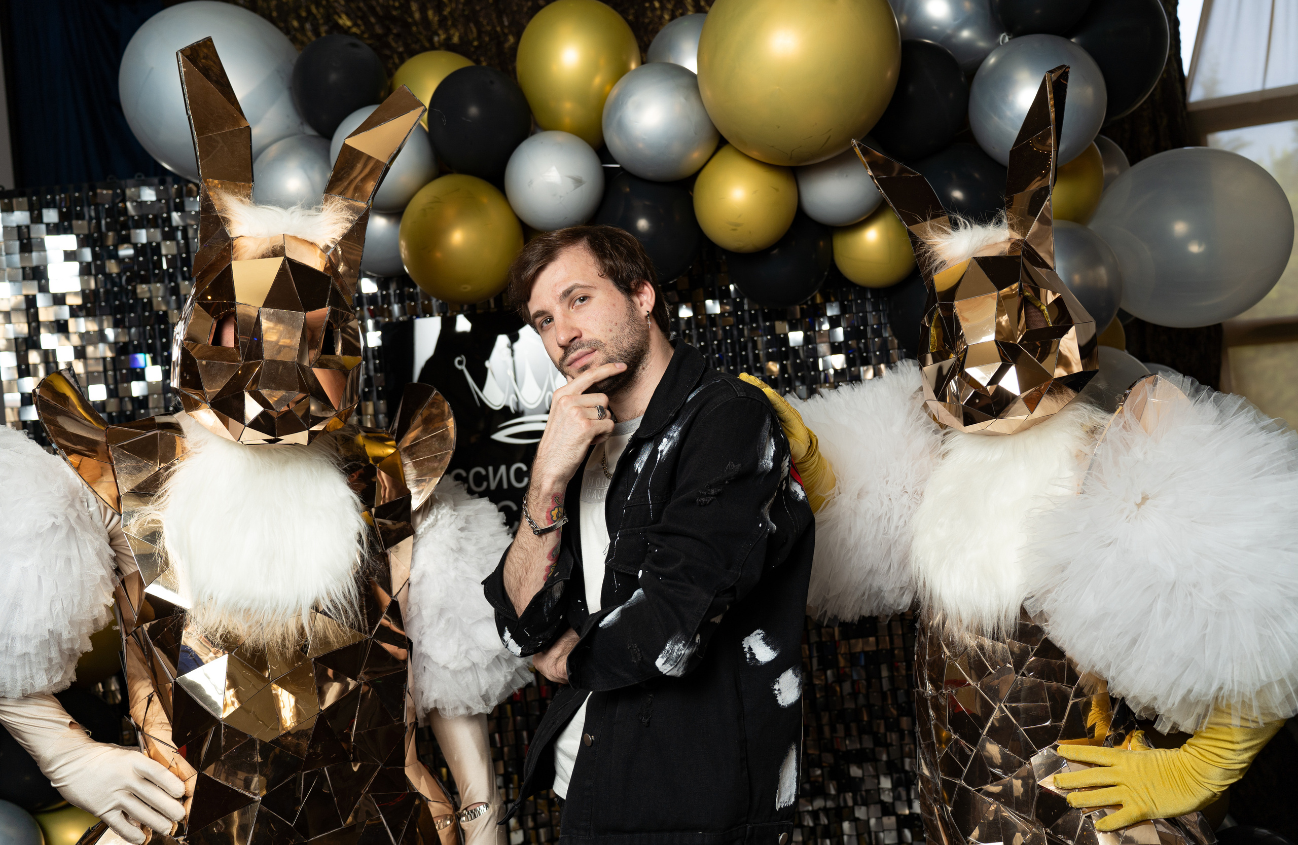 Это изображение мужчины, стоящего между двумя людьми в костюмах кроликов. Мужчина одет в черную куртку, джинсы и белую футболку. У него борода, а волосы короткие и темные. Он смотрит в камеру и улыбается. Люди в костюмах кроликов одеты в белые меховые костюмы с золотыми вставками. Они стоят по обе стороны от мужчины и тоже смотрят в камеру. На заднем плане много серебряных и золотых воздушных шаров.