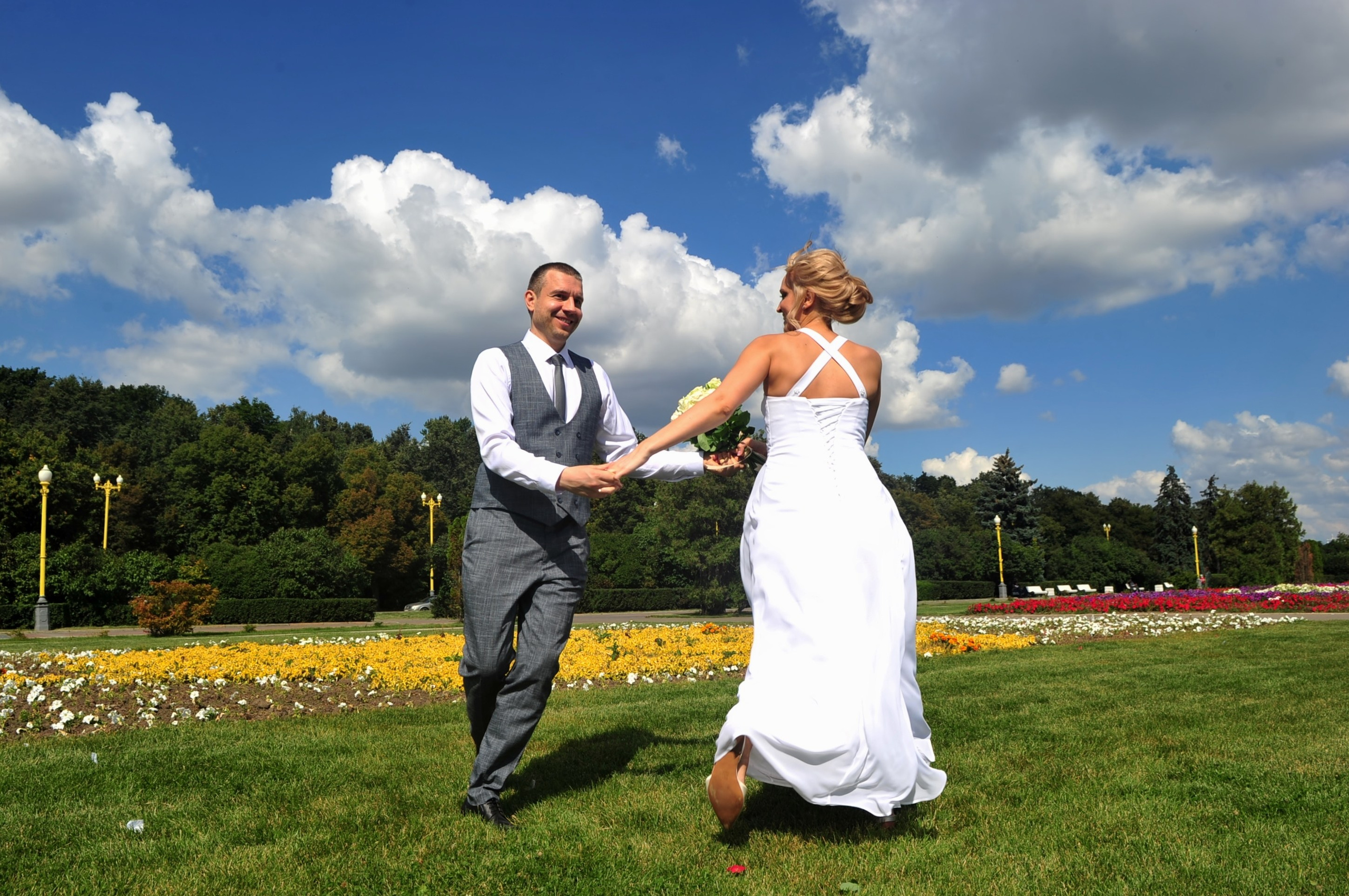 Свадьба на Воробьёвых Горах рядом с МГУ, новобрачные держатся за руки на фоне голубого неба, цветов и зеленой травы, жених улыбается невесте