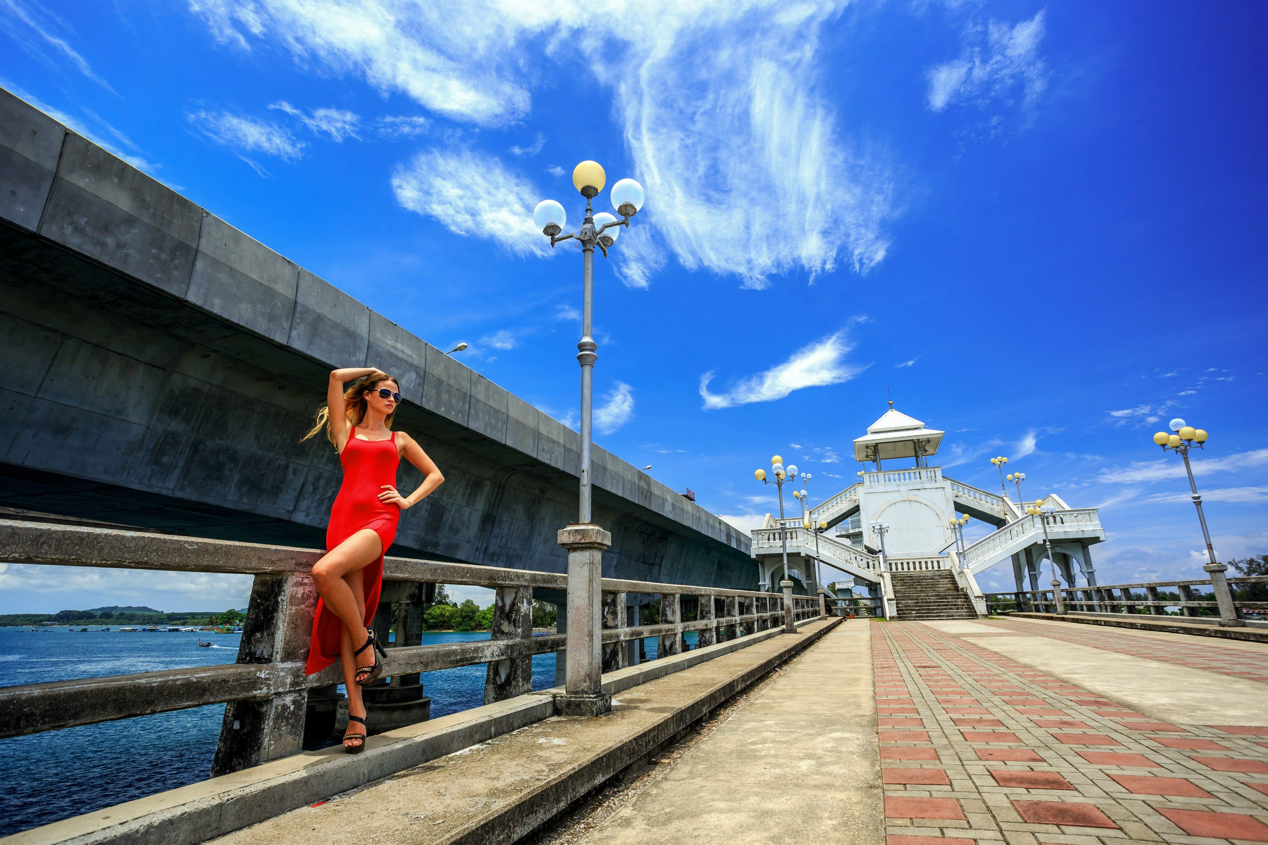 Fashion photo of a woman in a red dress posing on a riverside bridge walkway under a bright blue sky with dramatic clouds, modern urban architecture, street lamps and water in the background, stylish outdoor fashion shoot with strong perspective and summer atmosphere.