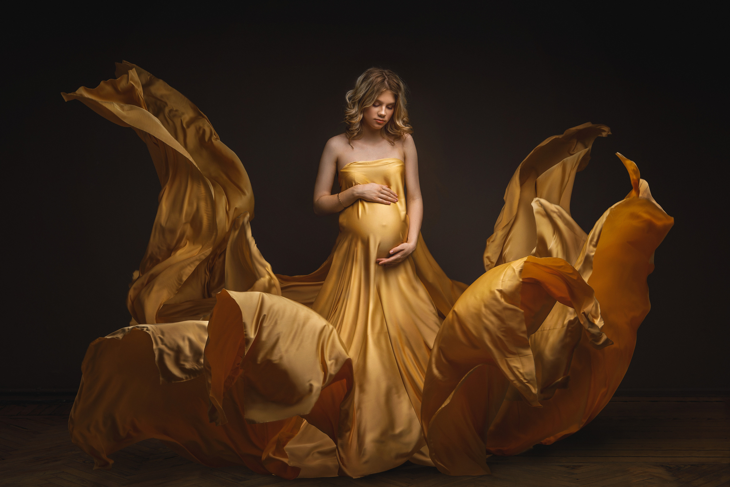 Беременная девушка позирует в фотостудии на темном фоне. На будущей маме красивая летящая золотая ткань 