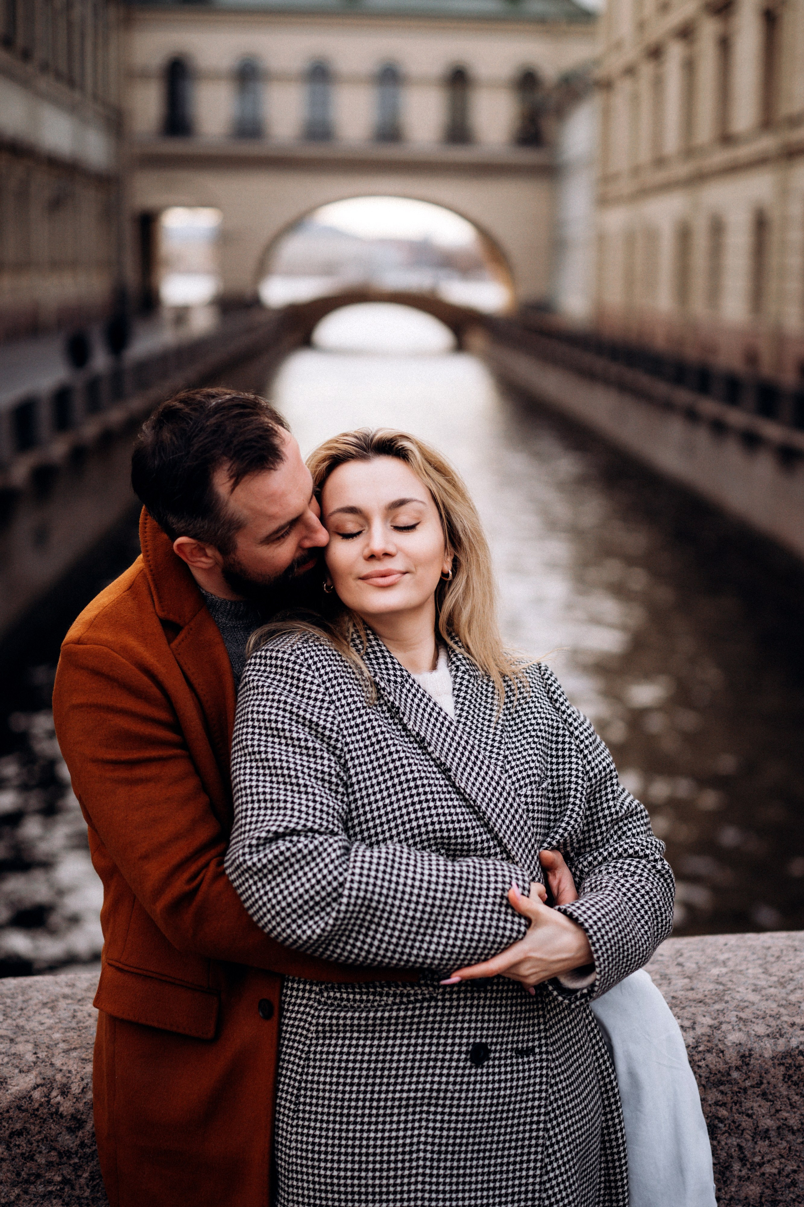 Лавстори Love Story в Санкт-Петербурге у Зимней канавки. Пара в  пальто обнимаются у гранитной ограды моста