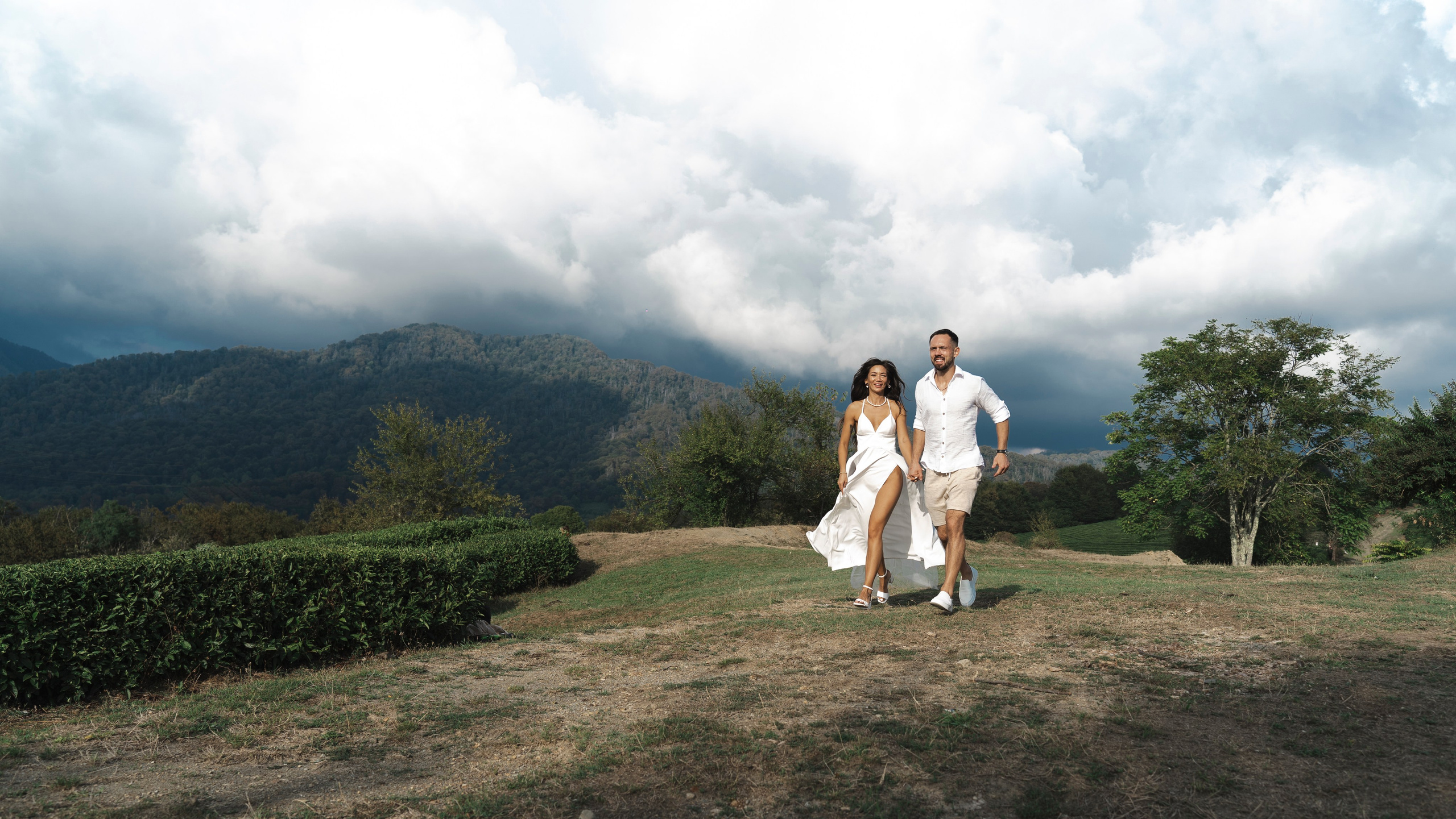 На фотографии изображен пейзаж с горами на заднем плане и облачным небом. В переднем плане видна травянистая зона с живой изгородью и несколькими отдельно стоящими деревьями. Местность кажется ухоженным полем или парком с грунтовой дорожкой, что указывает на естественную, открытую обстановку. По полю идут мужчина и женщина, держась за руки, оба одеты в белое. Женщина в воздушном платье, а мужчина в белой рубашке, шортах и кедах, что создает расслабленную, но элегантную атмосферу. Общий вид изображения подразумевает, что это момент из досугового мероприятия на открытом воздухе или фотосессии пары.