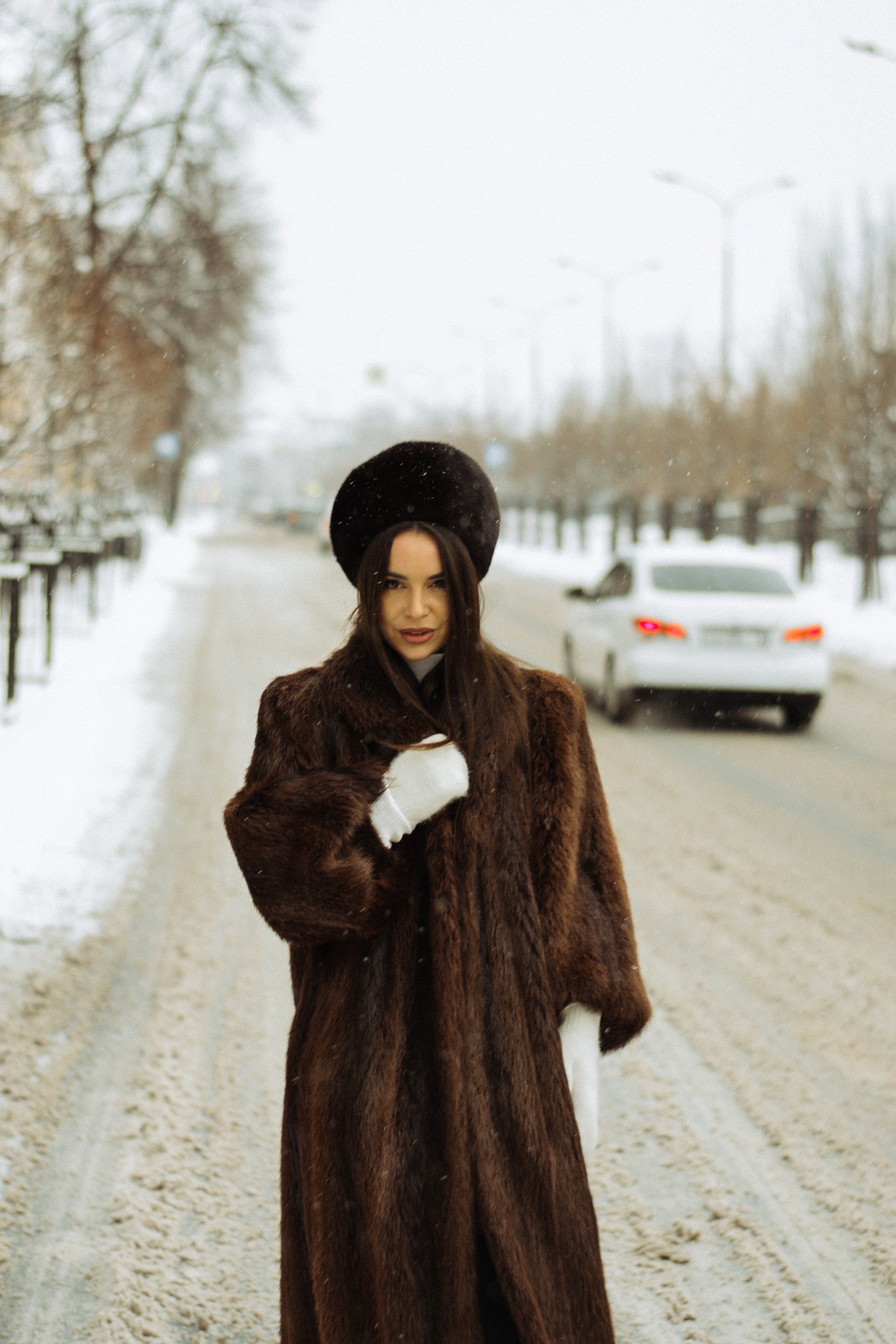 Женщина в коричневой шубе и меховой шапке стоит на заснеженной улице.