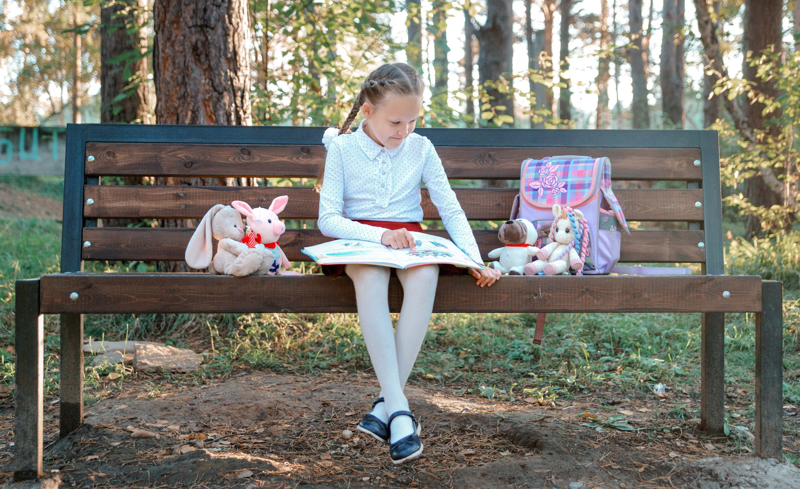 Детская фотосессия осенью - школьница сидя на лавочке читает книгу игрушкам