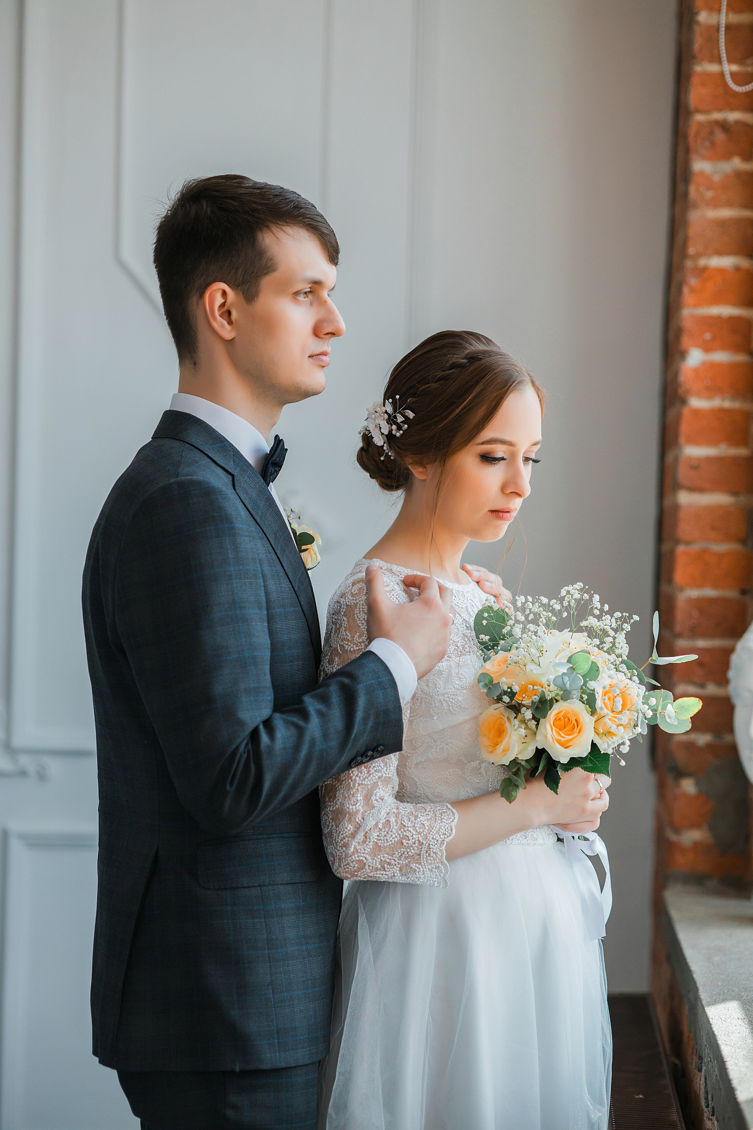 Свадебная пара обнимается. Девушка держит цветы. Пара стоит около окна.