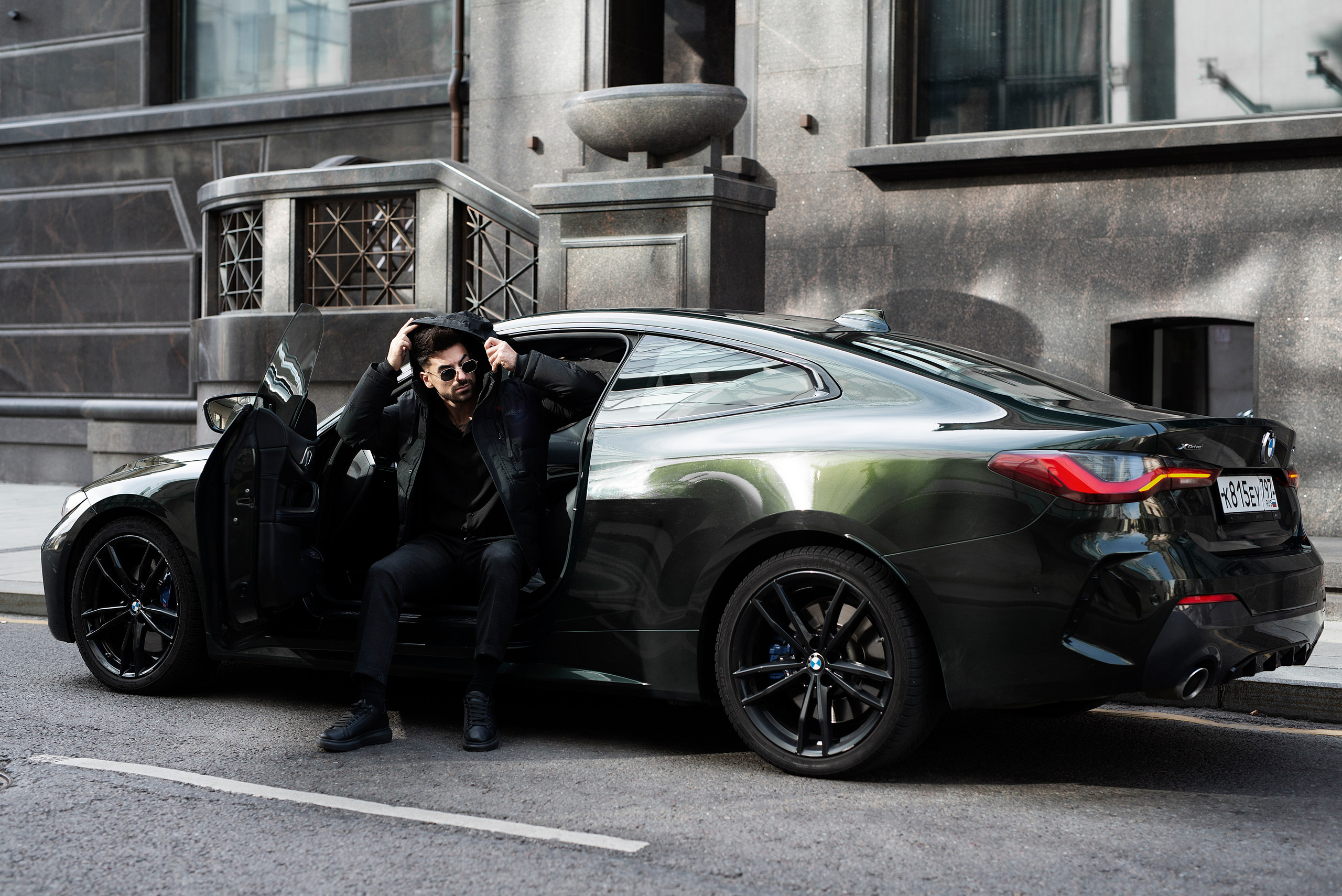 Брутальный и стильно одетый мужчина позирует сидя в BMW на фоне  городского пейзажа