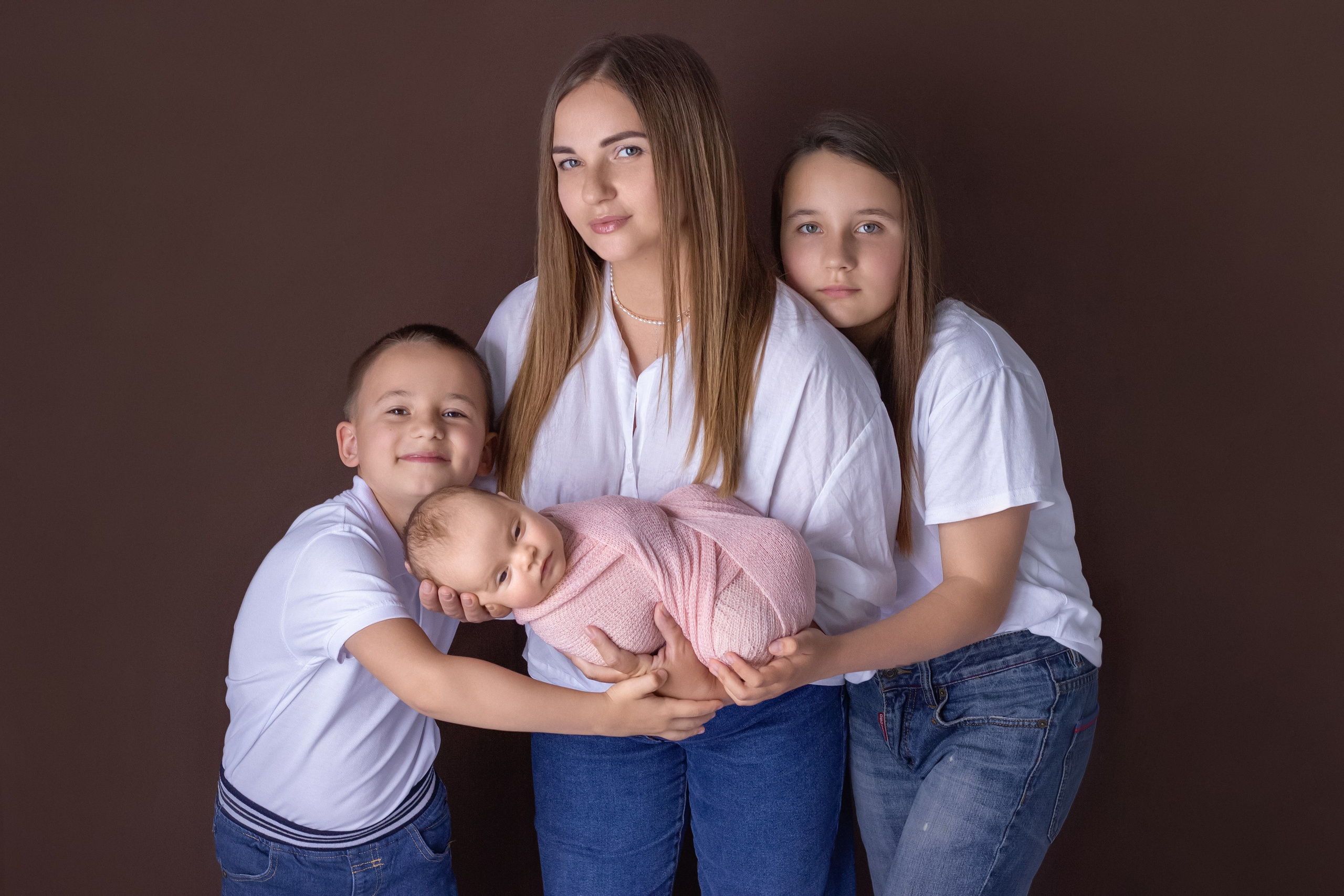 домашний портрет на коричневом фоне счастливой семьи с детьми и новорожденной, белые футболки и джинсы, все обнимаются и держат малышку