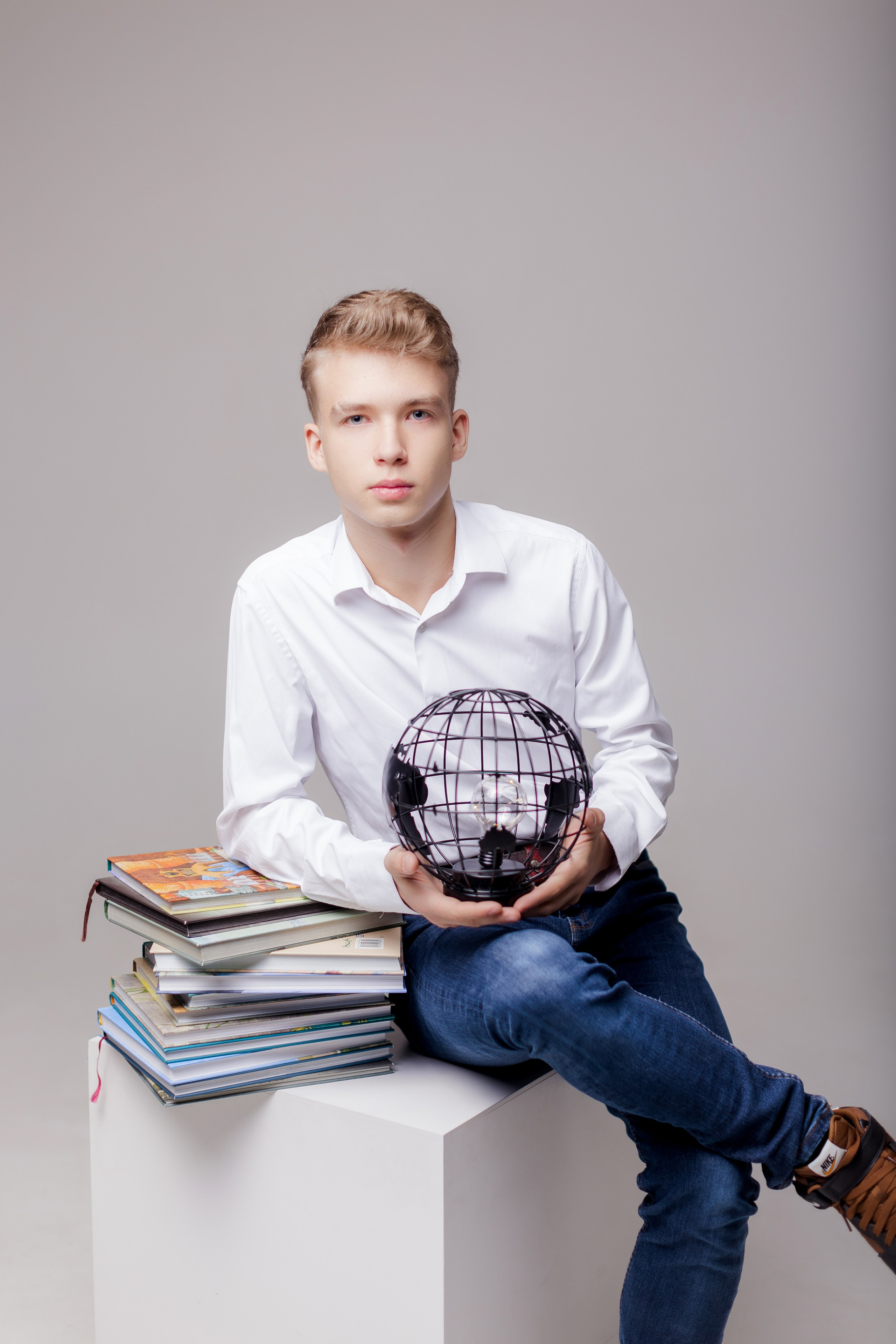 Молодой человек сидит в фотостудии на тумбе с книгами и глобусом