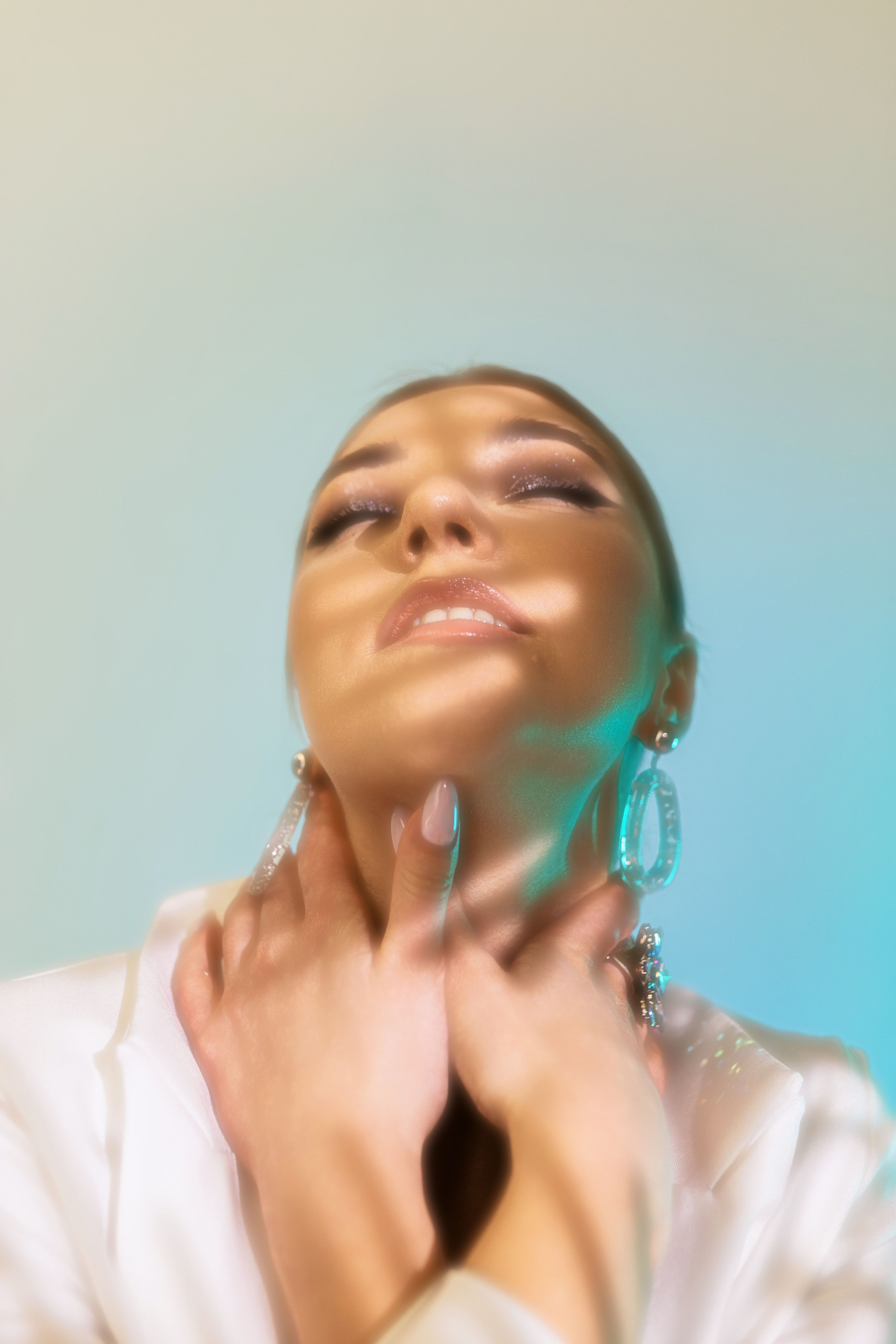 Уверенная в себе женщина с потрясающим макияжем демонстрирует свои элегантные серьги, нежно касаясь шеи, создавая ощущение гламура и утонченности в обстановке с мягким освещением.