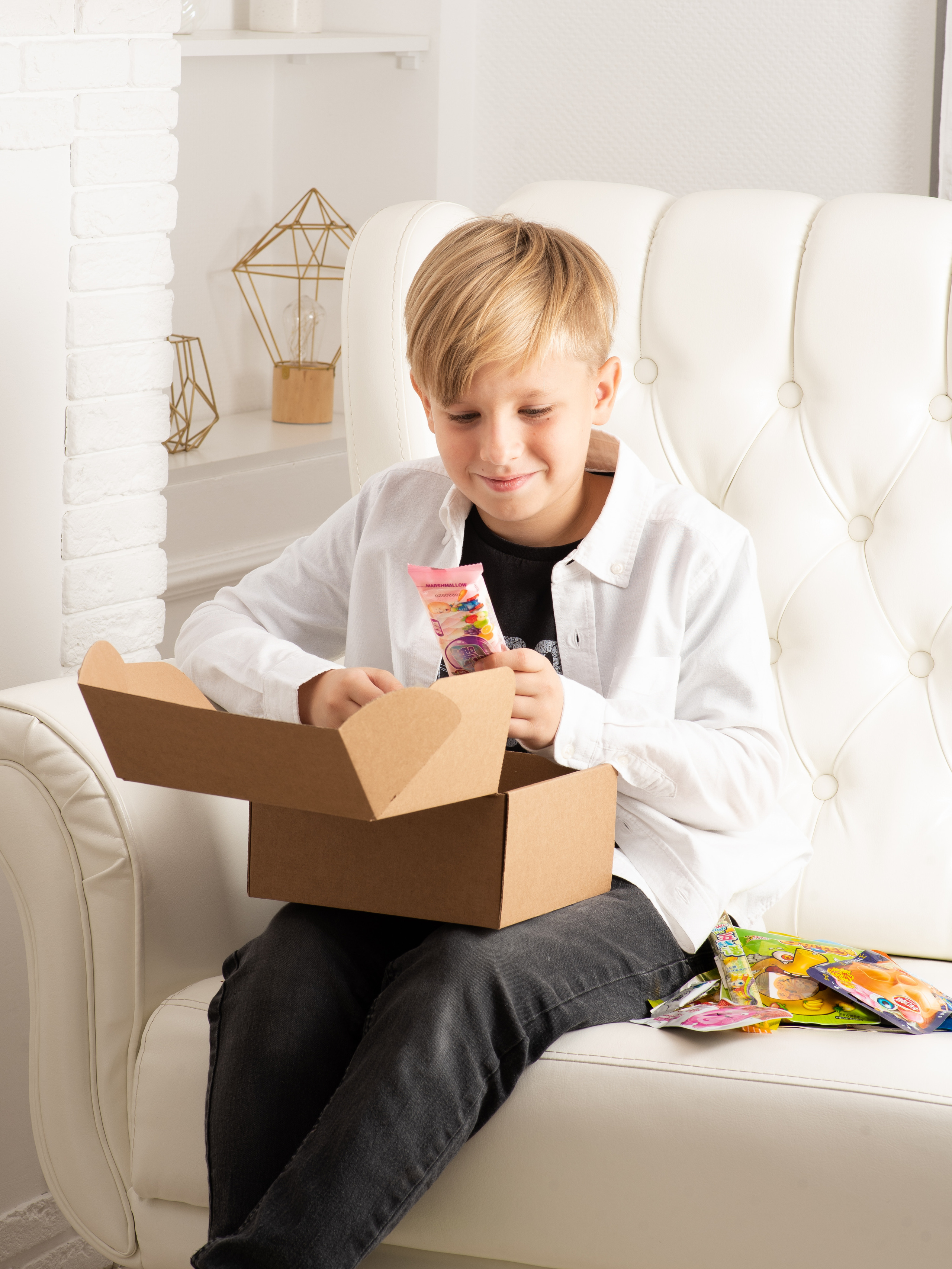 Симпатичный мальчик сидит на белом диване и ест конфеты. контент съемка.