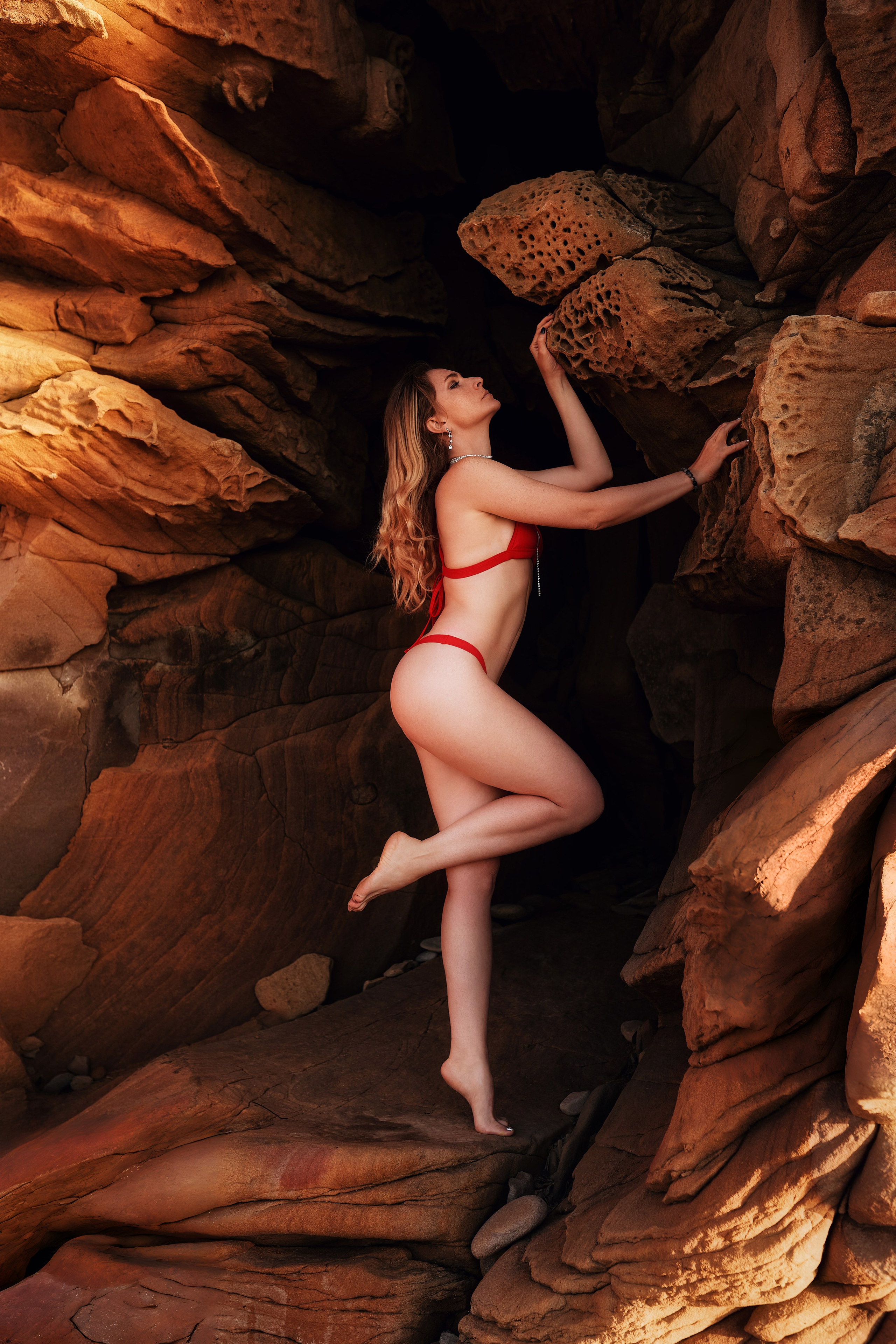 Зрелая модель в красном купальнике позирует на фоне фактурных прибрежных скал