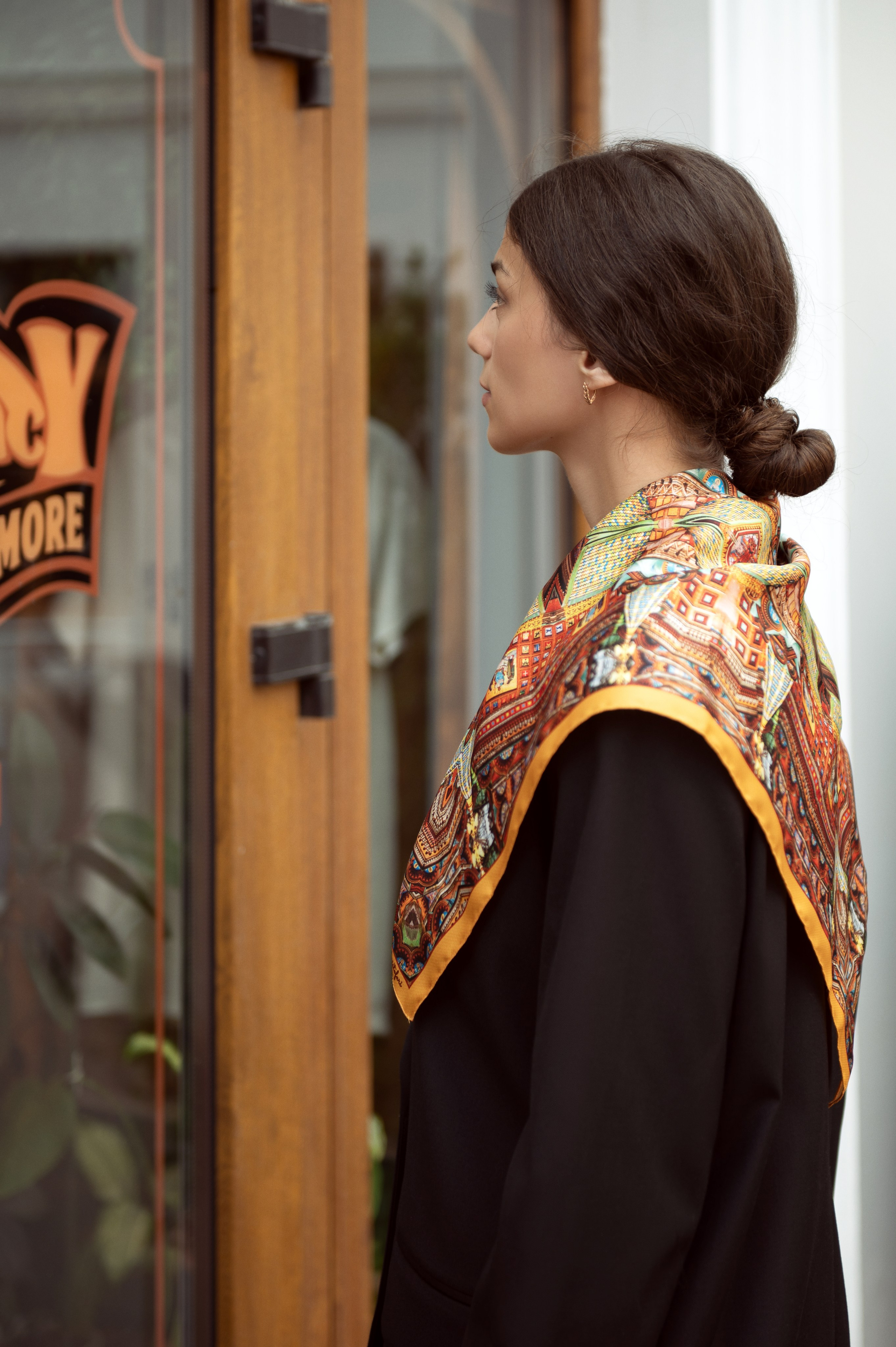 Красивая темноволосая девушка в черном пиджаке смотрится в витрину магазина, у нее на плечах шелковый платок от Serafim Cristofari