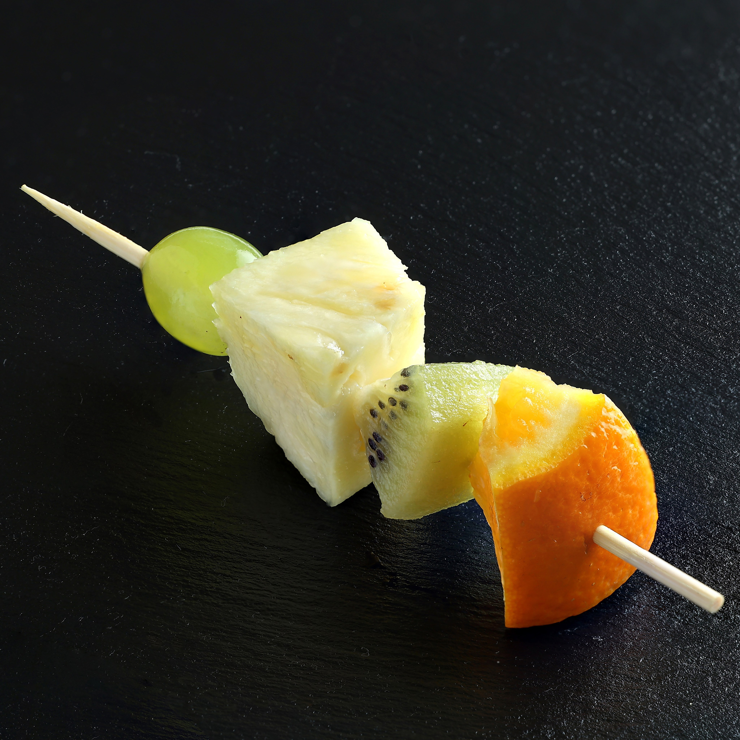 Фруктовая шпажка с долькой апельсина, киви, ананасом и белой виноградиной на длинной деревянной шпажке, на тёмном фоне.