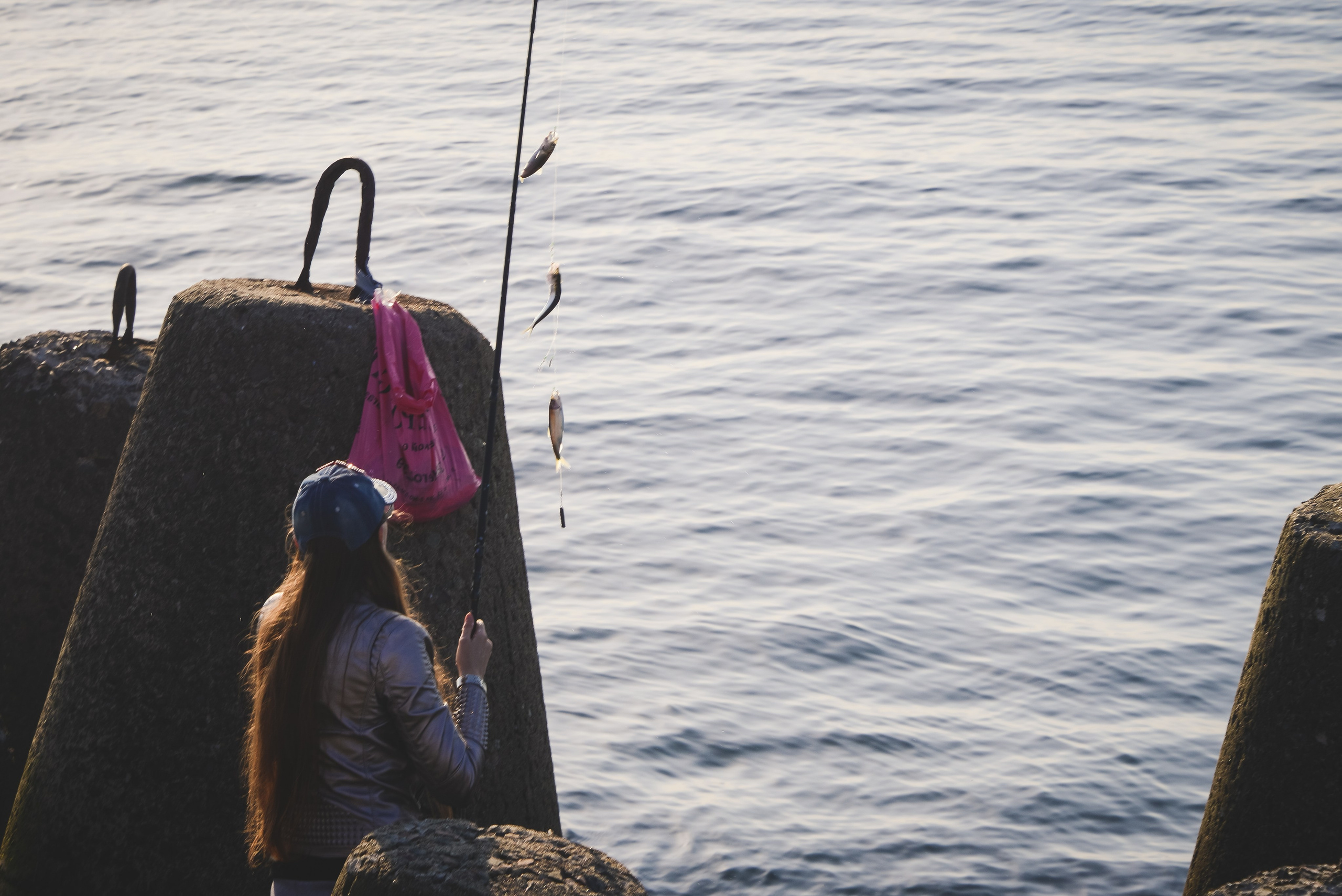 Девушка с длинными волосами в кепке ловит рыбу удочкой с рыбой на фоне моря. Рыбалка, хобби, отдых, досуг